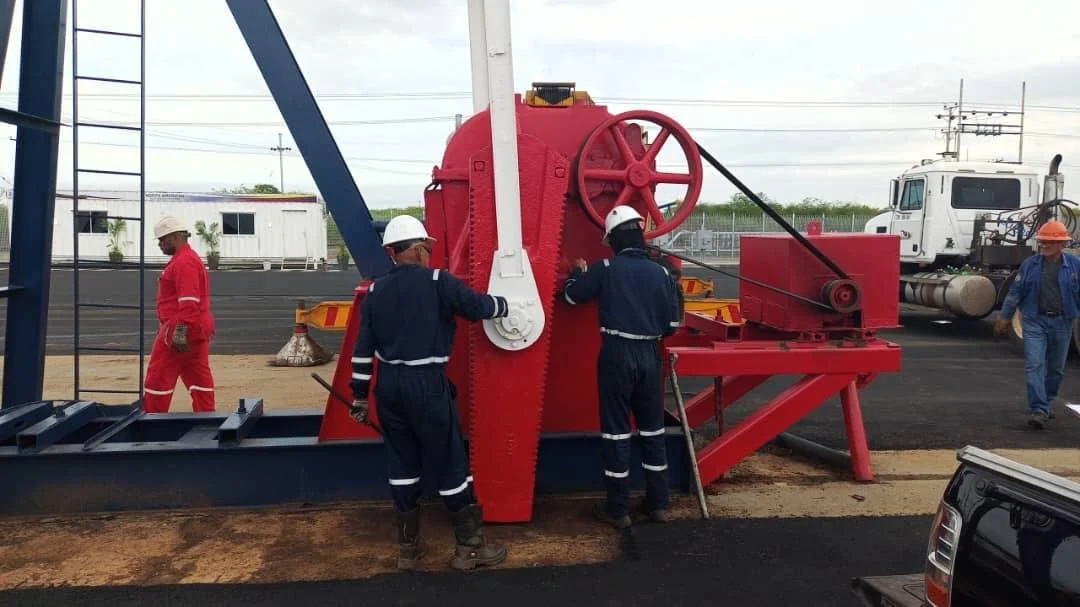 Workers in safety gear operating large heavy machinery on a construction site, with a truck and small buildings visible in the background.