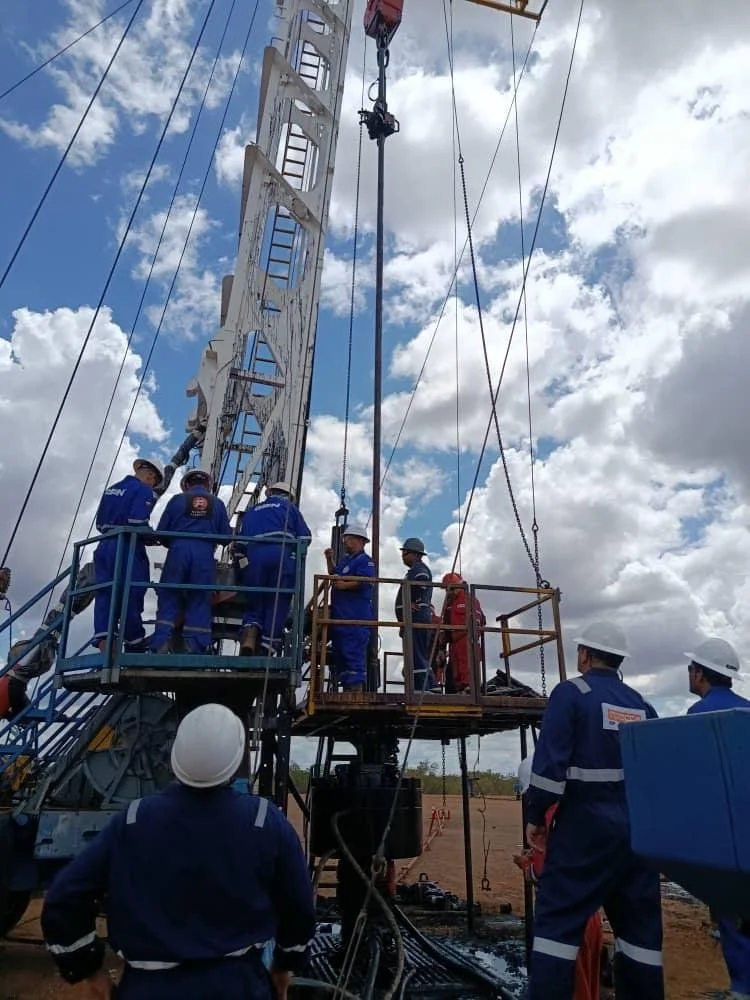 Workers in safety gear and uniforms operating a large oil drill rig outdoors under a partly cloudy sky.