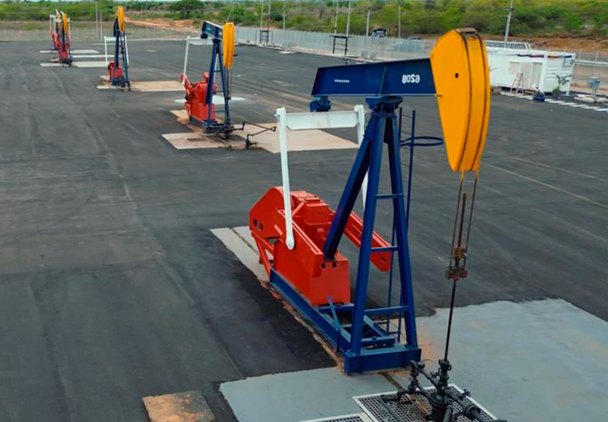 Multiple oil pumpjack machines on a large, empty, asphalt-covered lot, with a fence and greenery in the background.