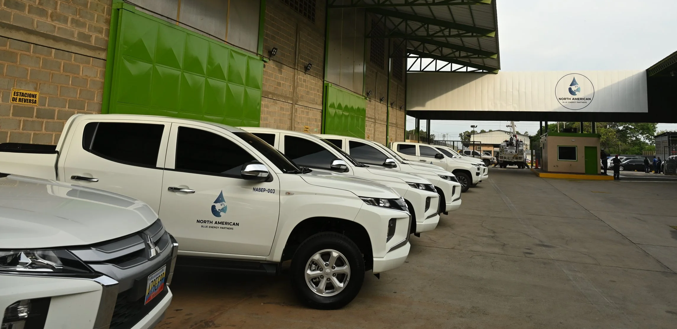Multiple white pickup trucks with North American Blue Energy Partners logos parked in a lot adjacent to a building. The area features green doors, a brick wall, and a large sign with the North American logo. Several people and a utility truck are visible in the background.