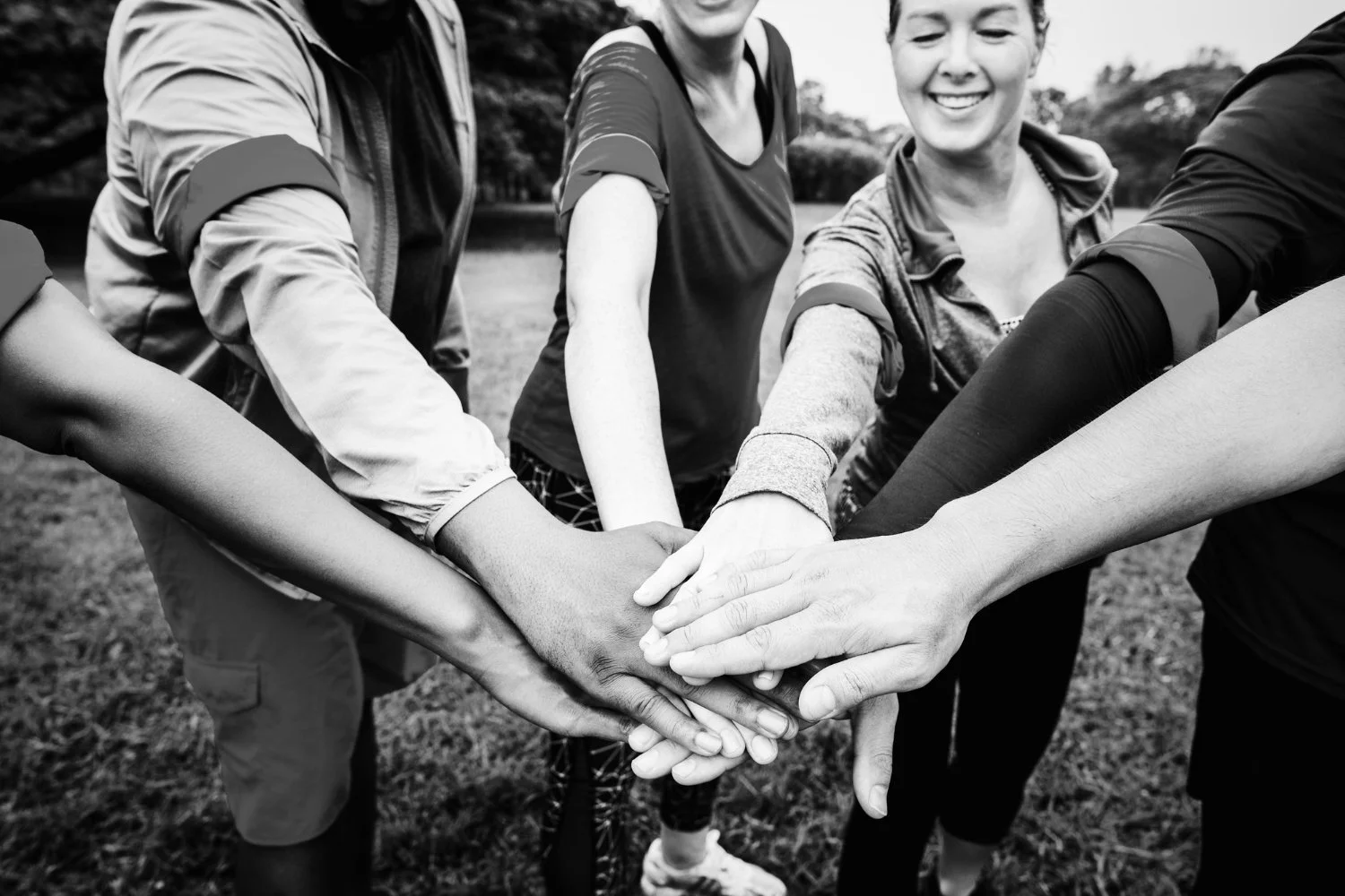 Group of diverse people in athletic clothing placing their hands together in a huddle outdoors, smiling and showing teamwork.