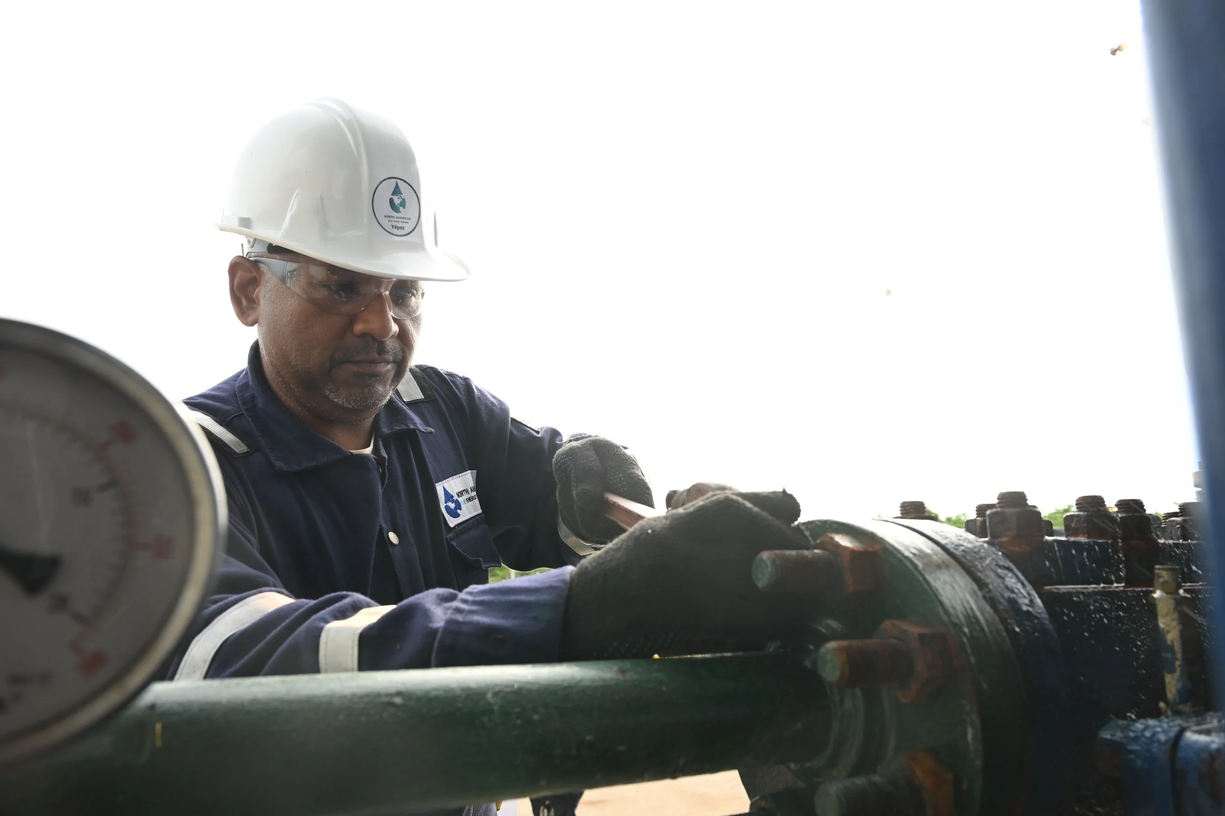 A man wearing safety glasses, a white safety helmet, and gloves inspecting industrial pipes and a pressure gauge outdoors.