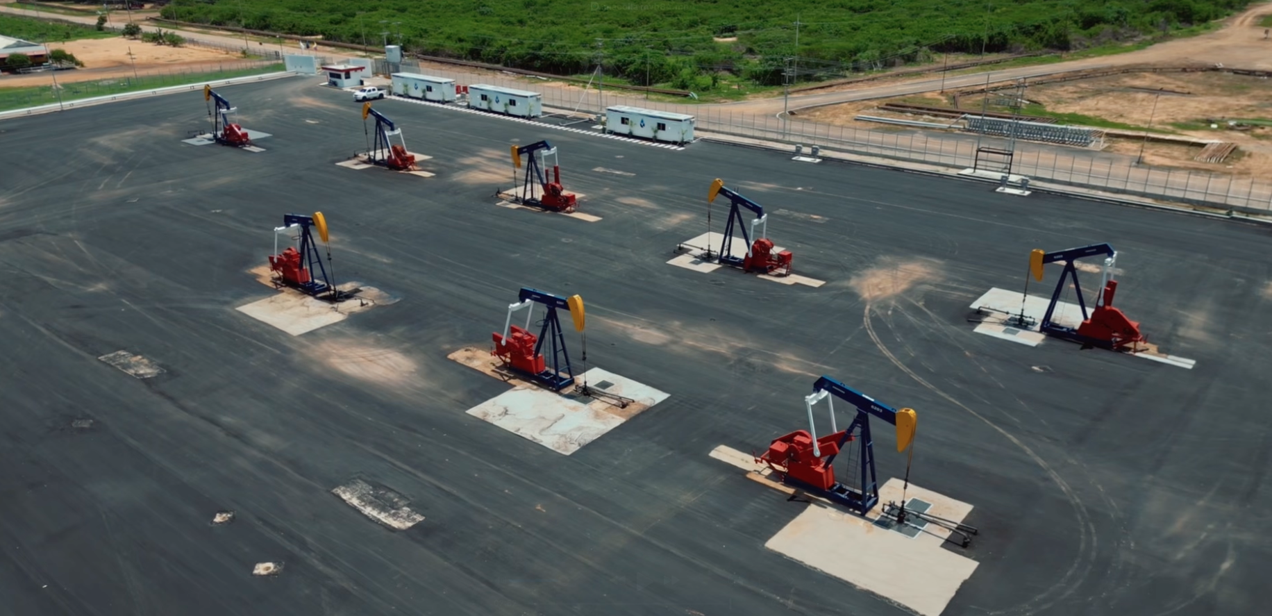 An aerial view of a large parking lot where seven oil pump jacks are installed in a grid formation, with some dirt patches visible around them and a few vehicles and small structures along the perimeter.