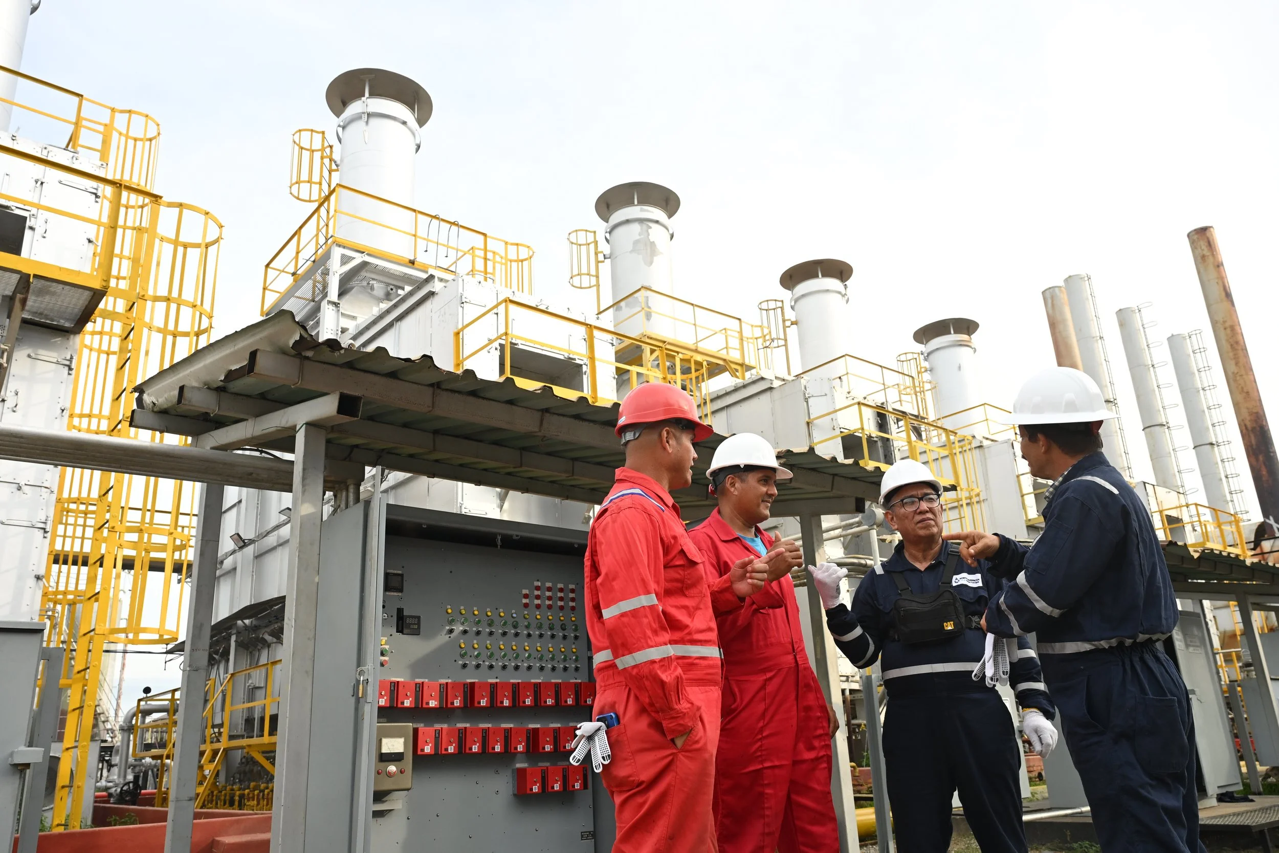 Four workers in safety gear, including helmets and coveralls, having a discussion at an industrial plant with equipment and pipes in the background.