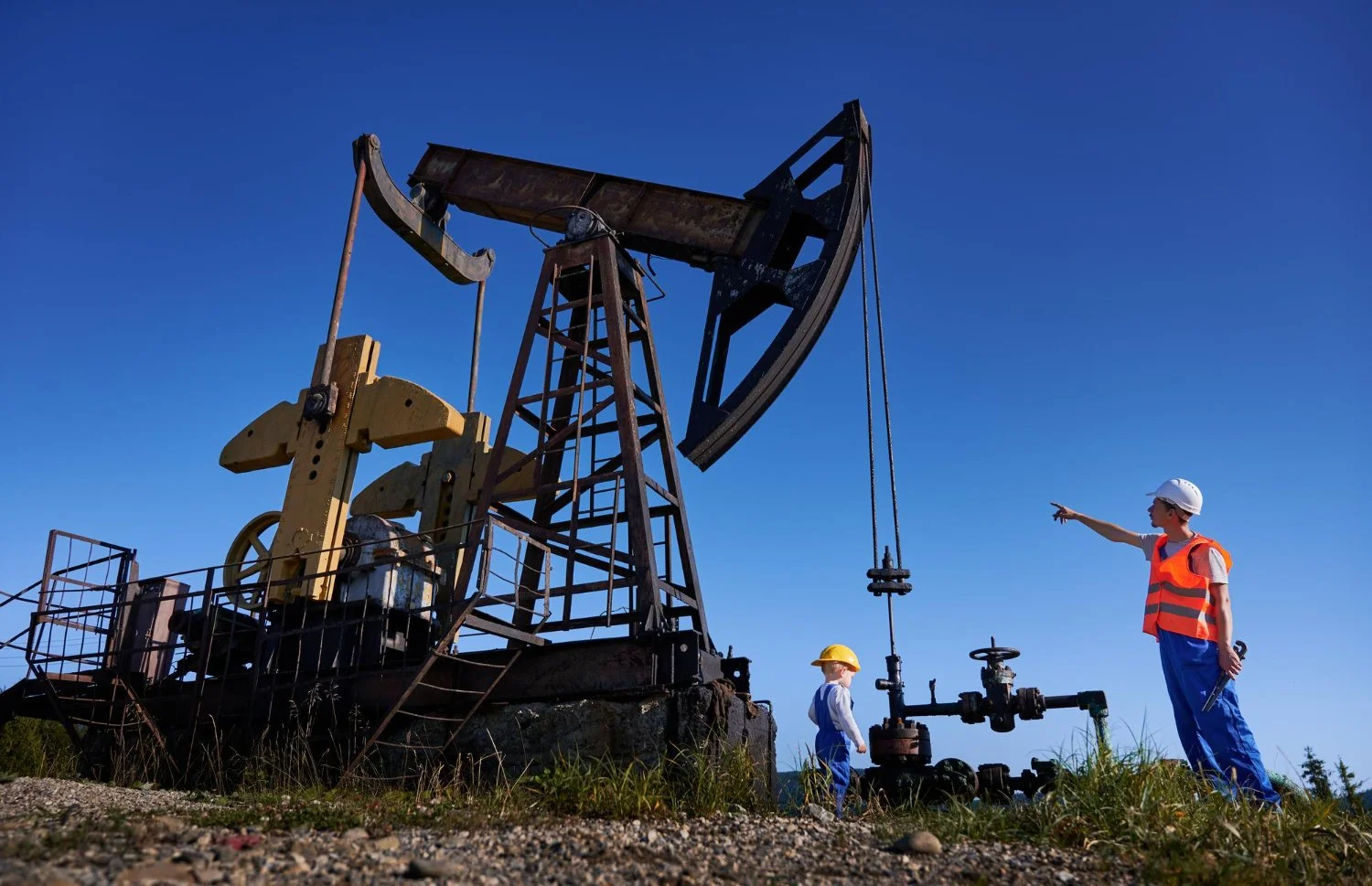 Two children dressed as oil rig workers standing next to a large oil pump jack outdoors under a clear blue sky, with the older child pointing.