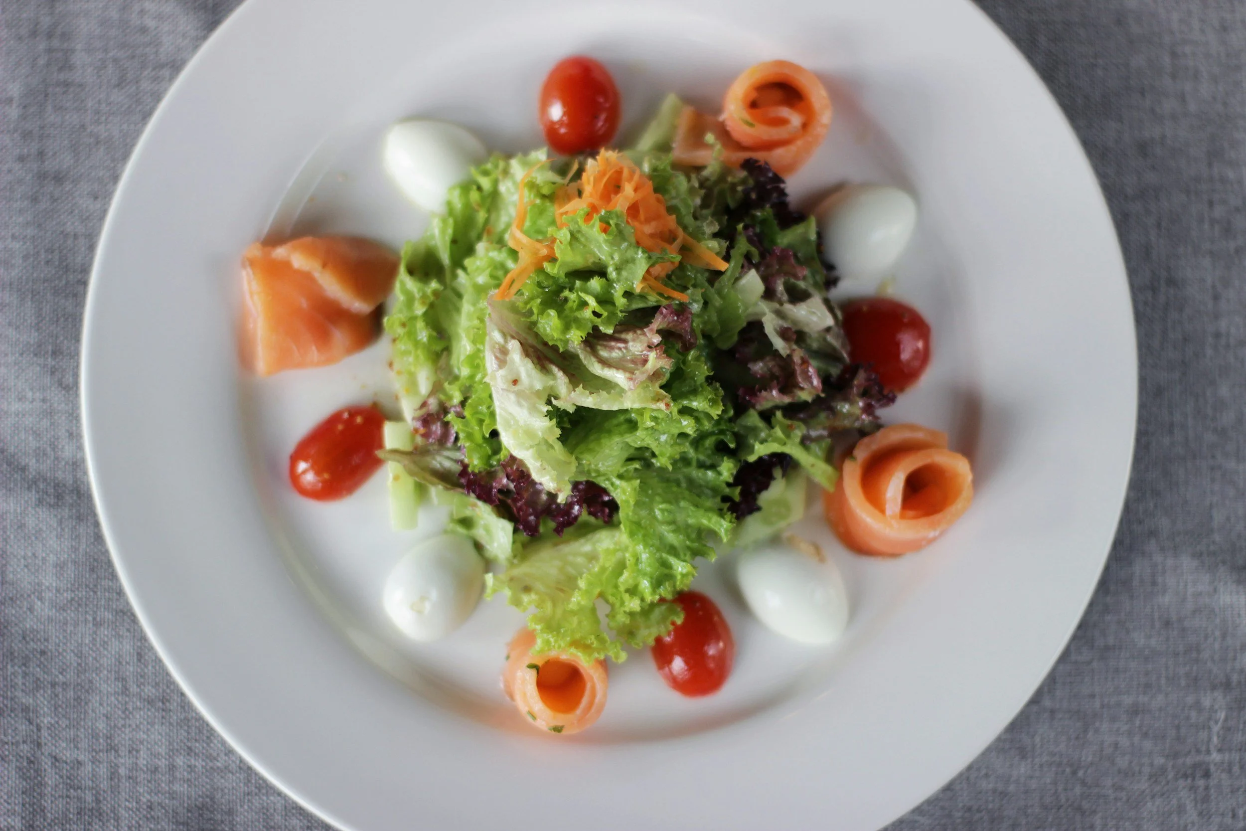 Mixed green salad with cherry tomatoes, rolled salmon slices, mozzarella balls, shredded carrots, and dollops of creamy dressing on white plate