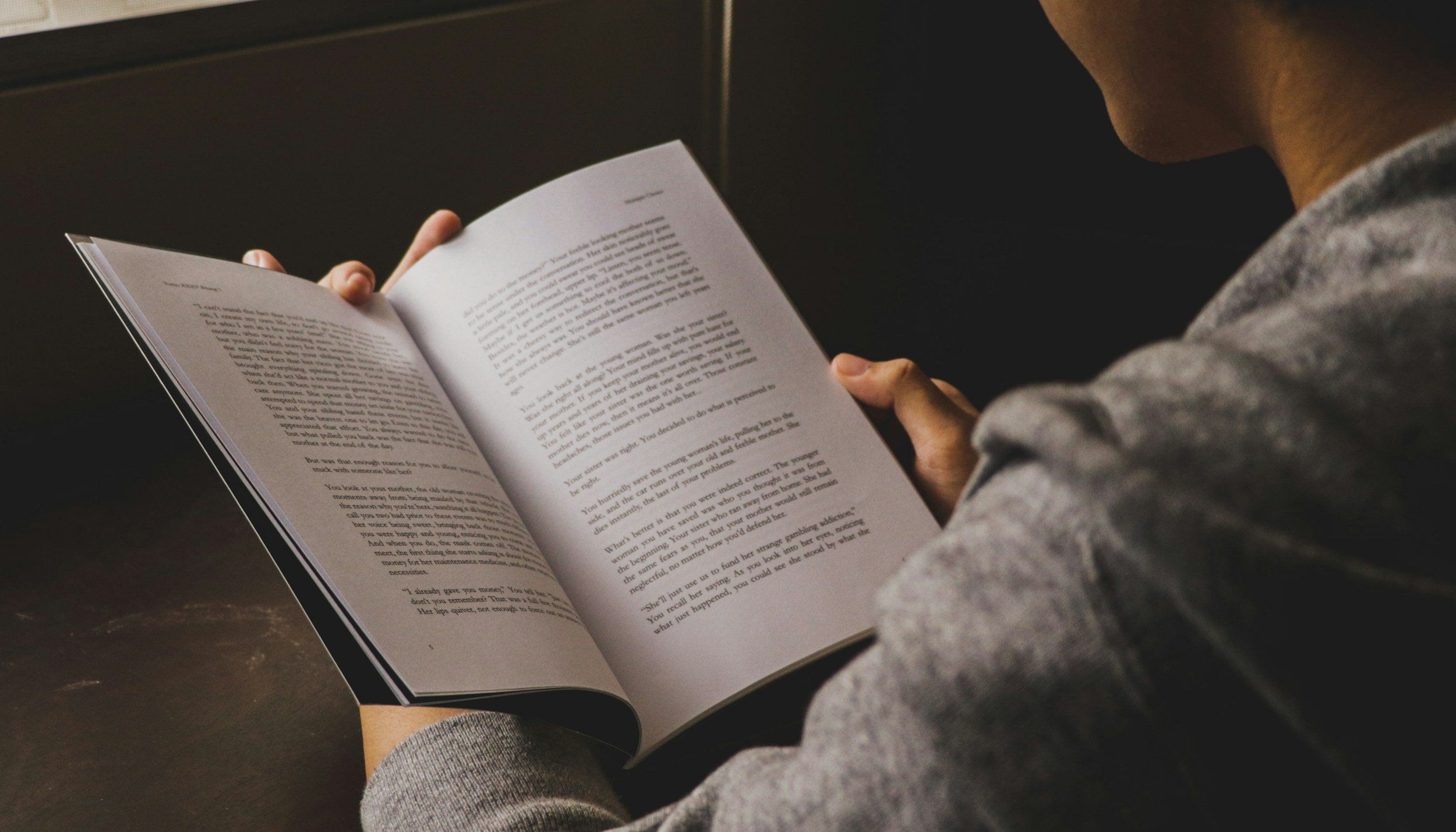 Person reading a book at a dark table, wearing a gray long-sleeve shirt.