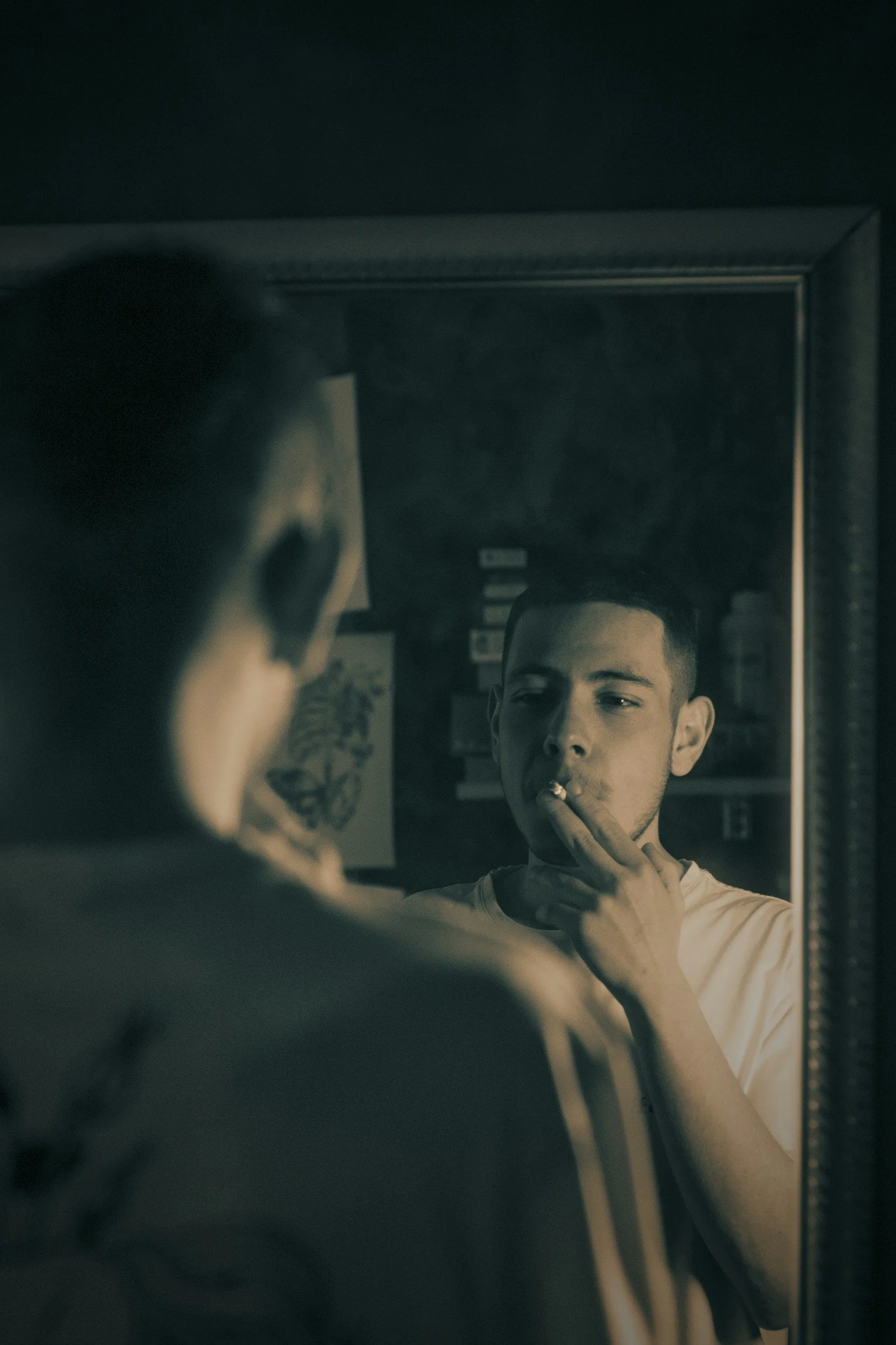 Young man looking at himself in a mirror, adjusting his chin while smoking a cigarette.