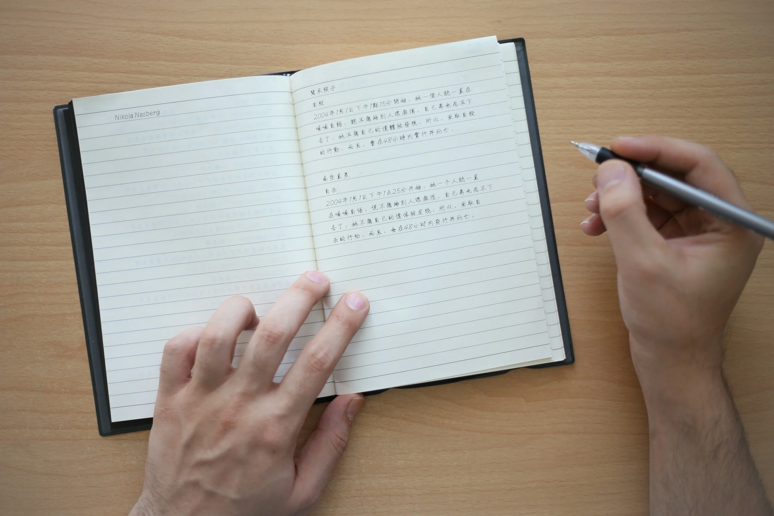 Open notebook on wooden desk with handwritten notes in Chinese, person's left hand holding down the left page, right hand holding a silver pen.