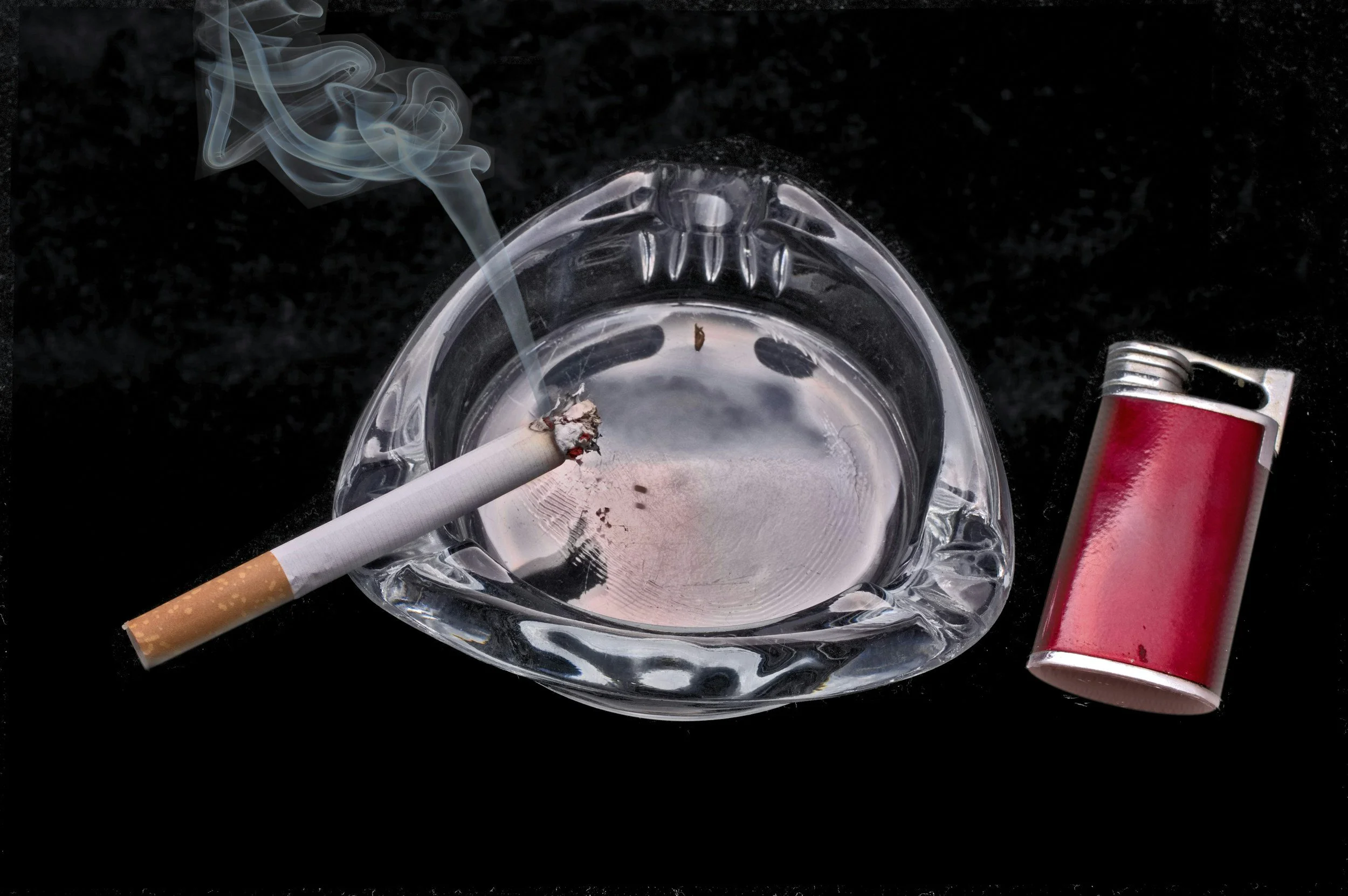 A lit cigarette with ash and smoke in a glass ashtray, a red disposable lighter, and a burning cigarette with smoke rising on a black surface.