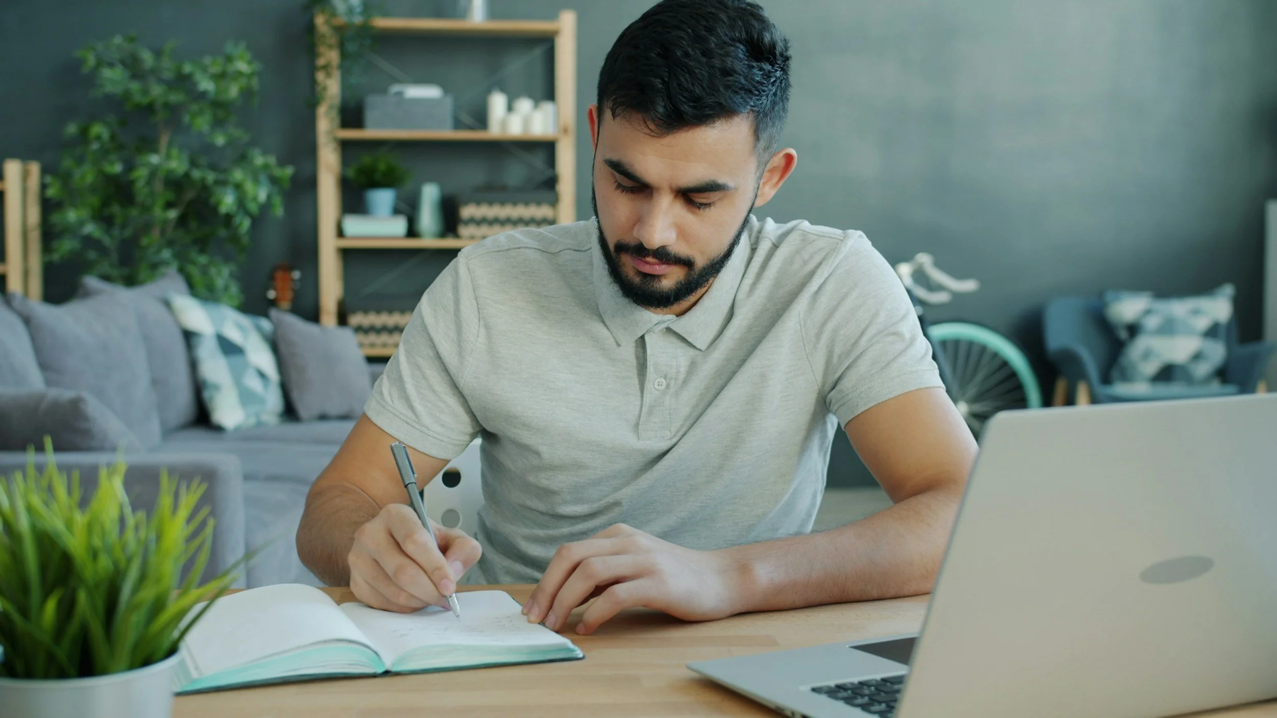 A young man with dark hair and a beard, wearing a light grey polo shirt, sitting at a desk writing notes in an open notebook with a pen. In front of him is a silver laptop, and to the left, a plant. The background features a black wall with wooden shelves containing decorative items and plants, and a blue bicycle partially visible.