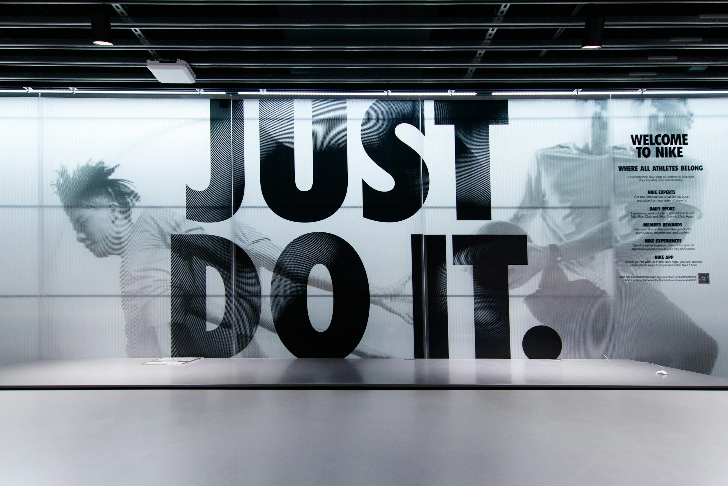 Store window display with large black text spelling 'JUST DO IT' and promotional information about Nike on the right side, with a person running in a black and white photograph in the background.