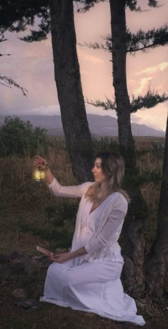 A woman in a white dress kneels outdoors near trees during sunset, holding a lantern in her right hand and a phone in her left.