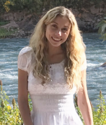 Young woman with long blonde hair smiling outdoors near water with rocks and greenery in the background.