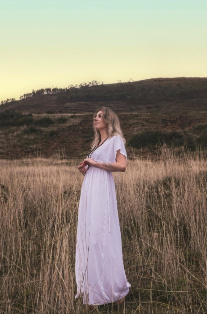 A woman standing in tall grass in a hilly, natural landscape during sunset or dusk, wearing a long white dress and looking to the side.