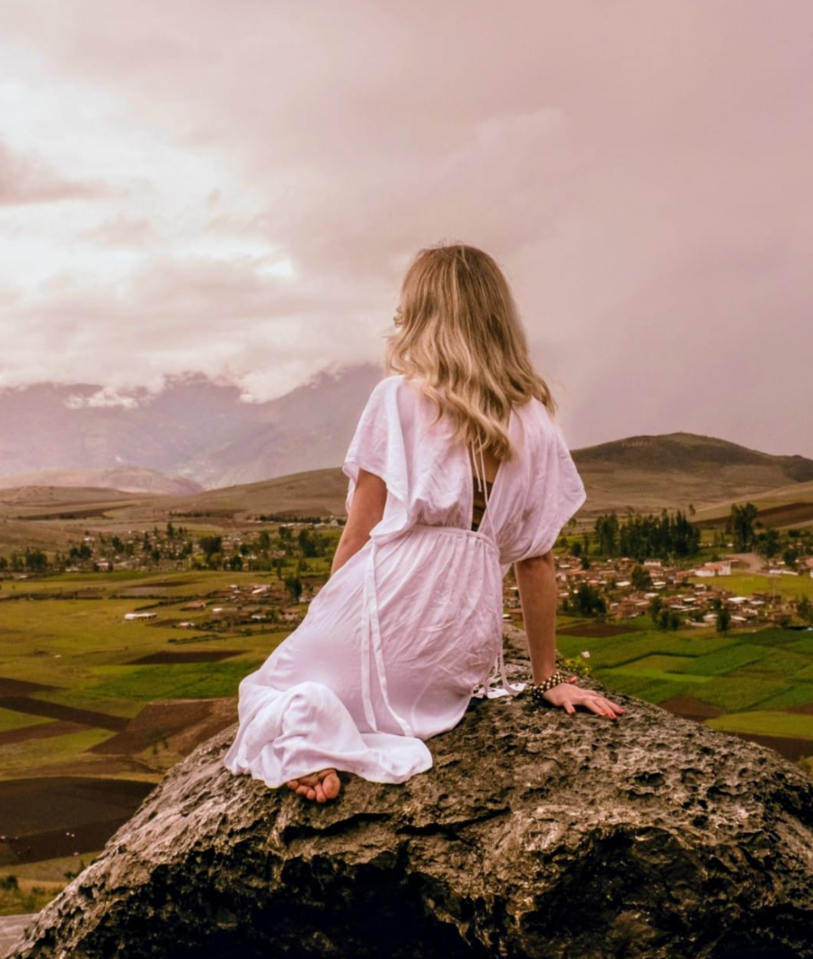 A woman dressed in white kneeling on a rock, overlooking a landscape of fields and distant mountains.