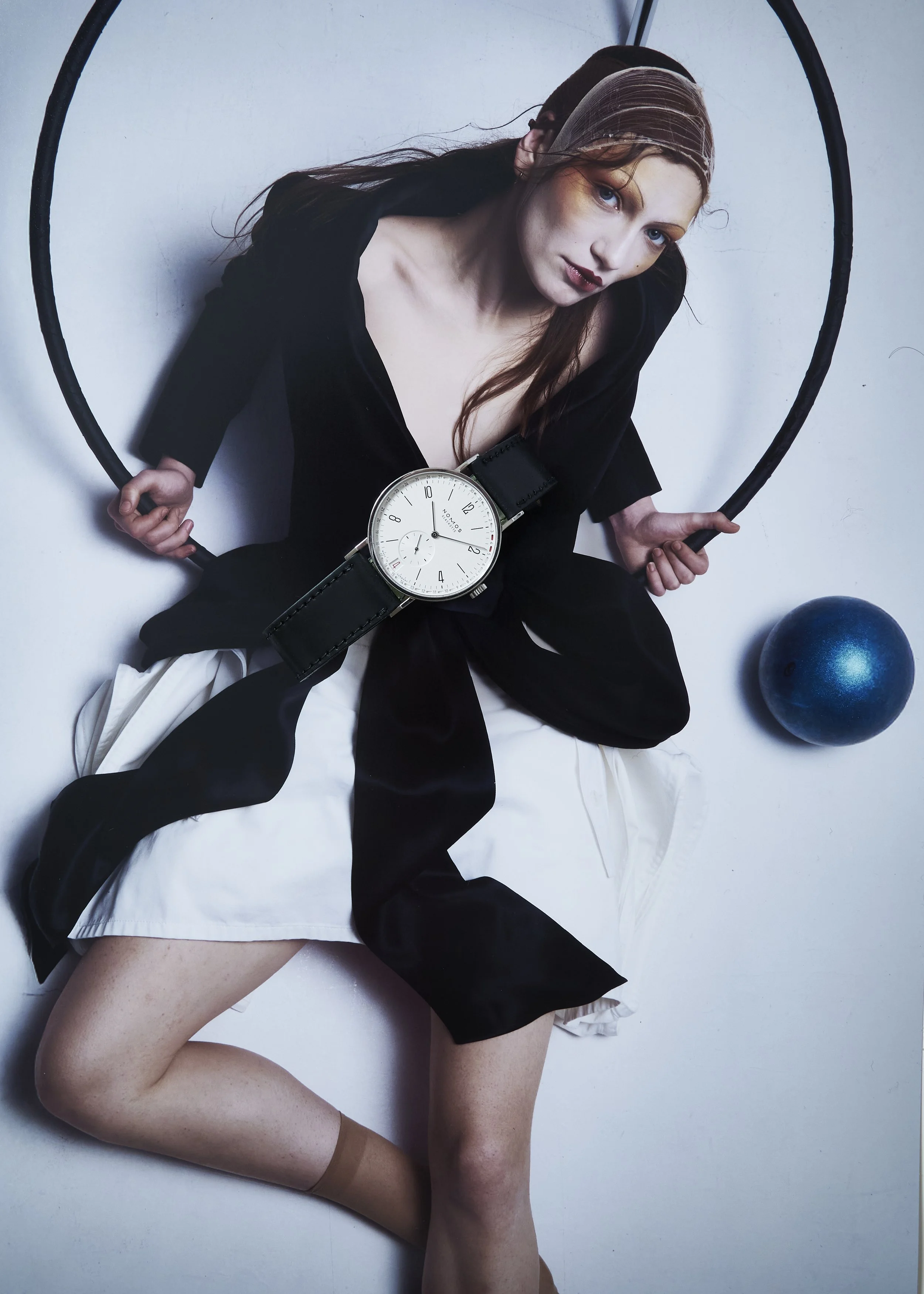 Femme avec un grand montre noire ayant un fond blanc, posant sur un sol blanc, avec une balle bleue à côté d'elle, tenant une barre noire.