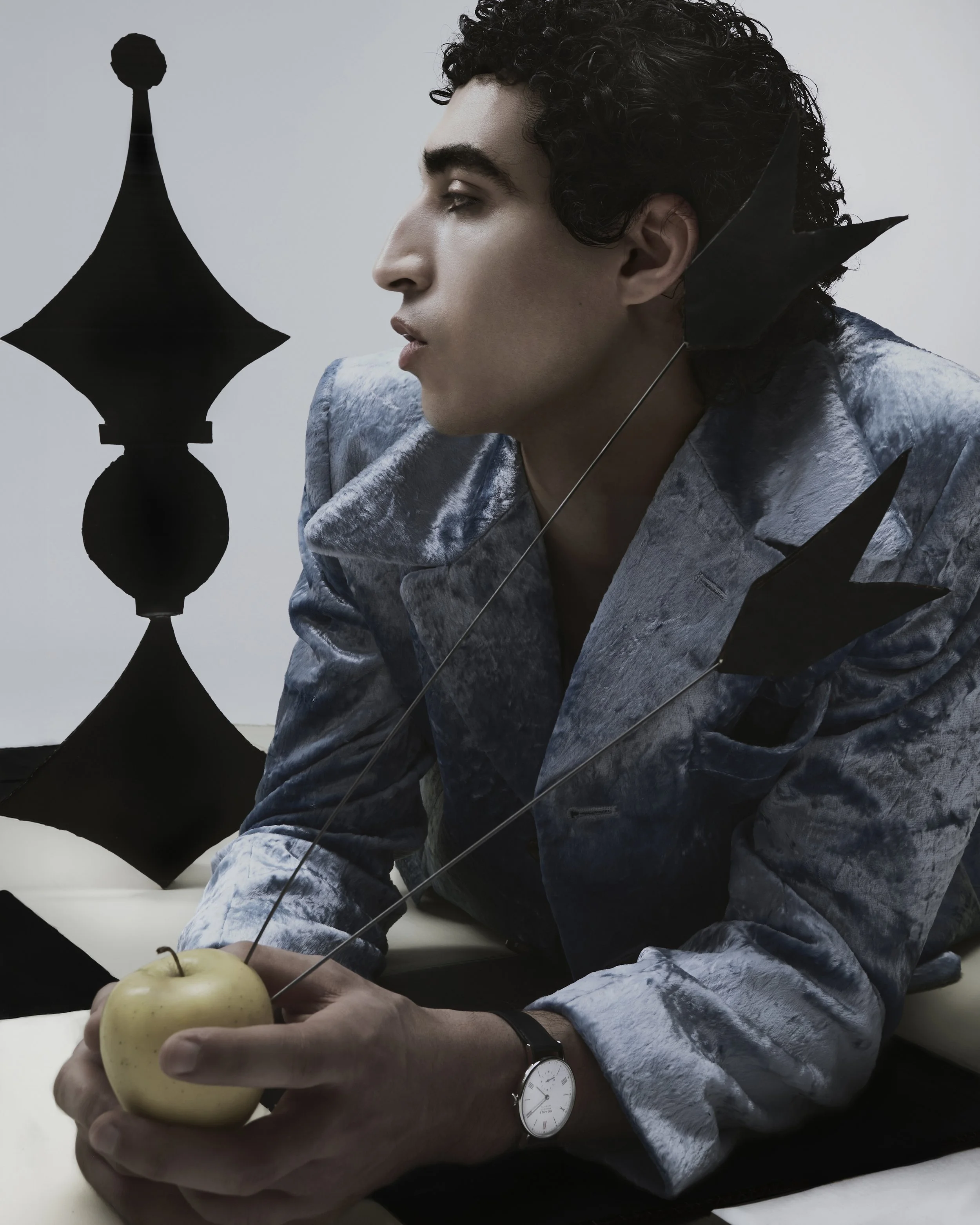 Un homme avec des cheveux bouclés porte un costume en velours gris et tient une pomme verte dans la main. Il porte une montre blanche. La photo est artistique avec des éléments de design noir en forme de flèches ou d'étoiles suspendues autour de lui.