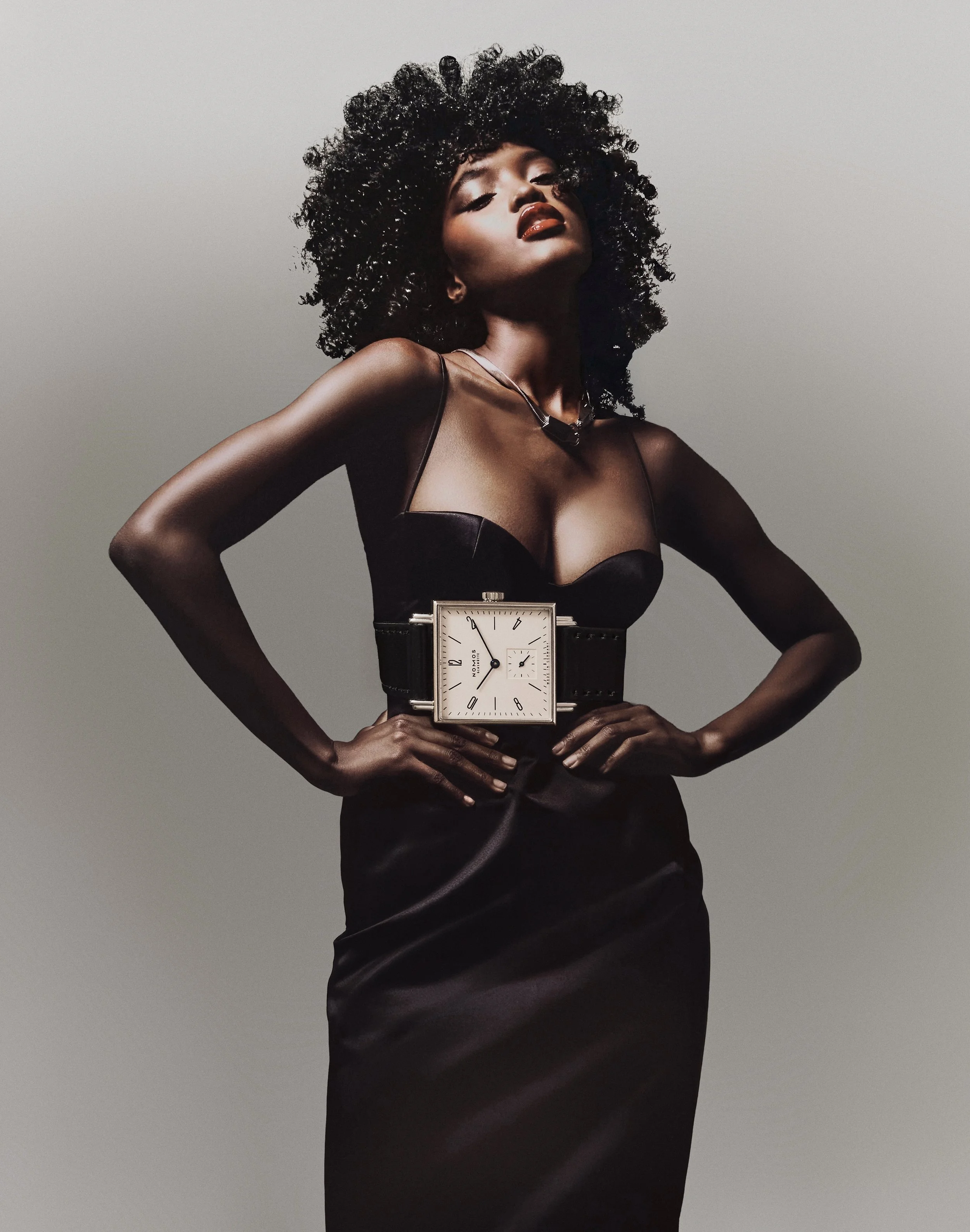 Femme noire élégante portant une robe noire et un gros Fat Watch sur la taille, avec cheveux bouclés et poses de confiance.