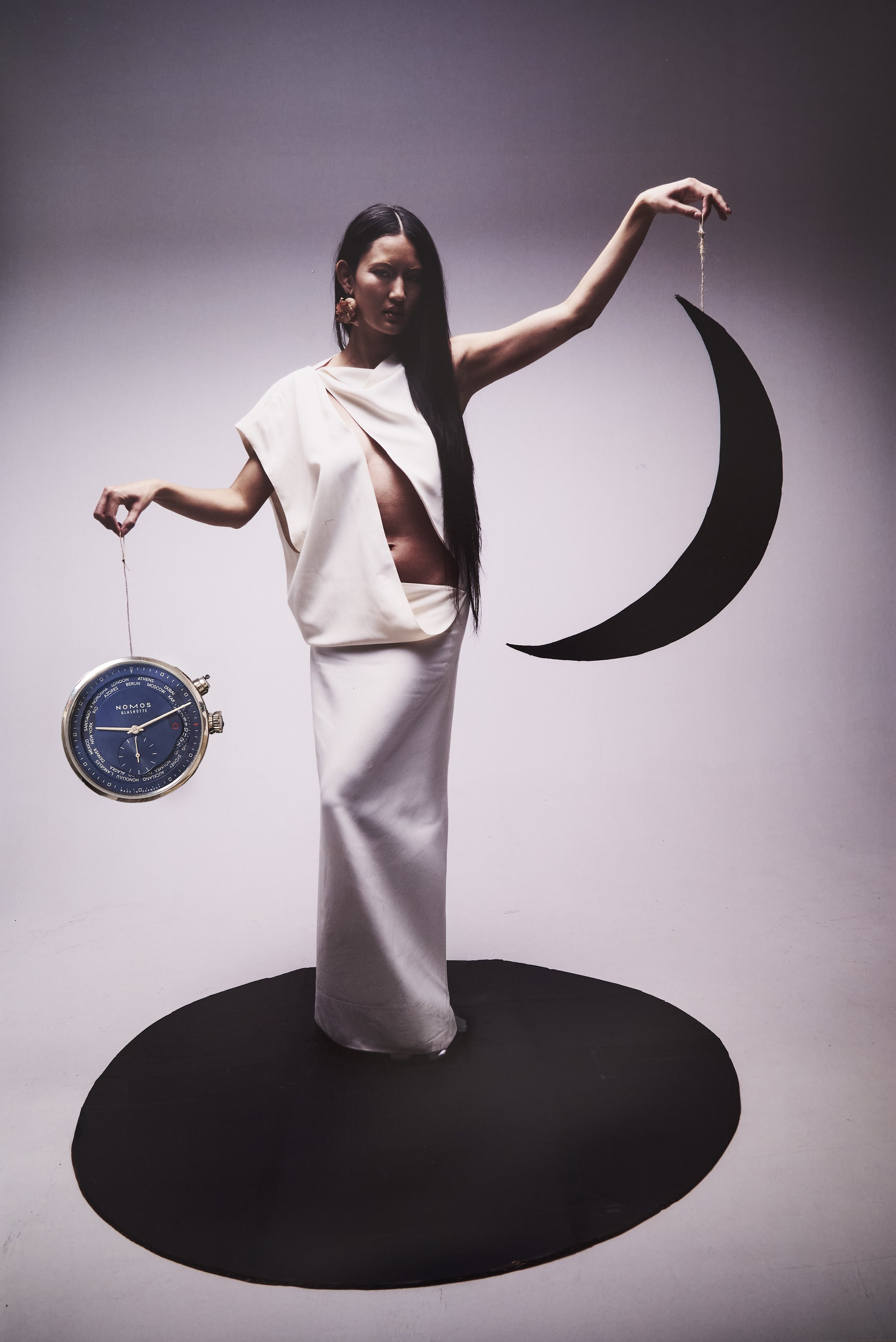 Une femme vêtu d'une robe blanche, posant sur un fond blanc, tenant une horloge et une lune en carton noir, créant une composition artistique.