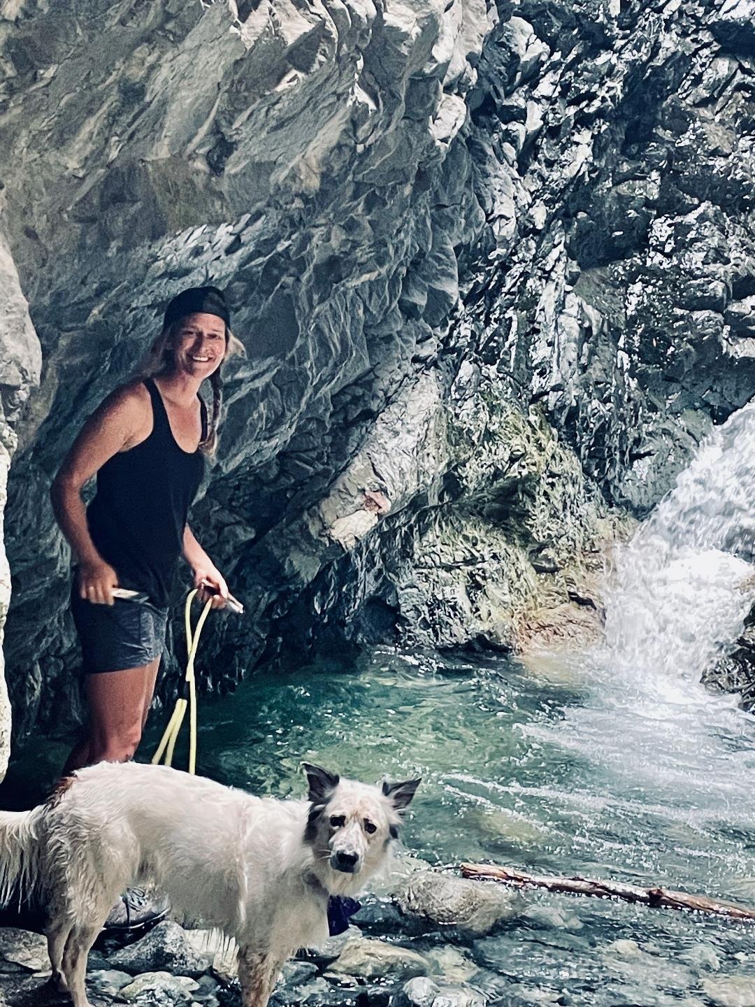 A woman with long hair, wearing a black tank top, shorts, and a black cap, standing next to a white dog with black markings and a bow tie, beside a rocky water stream, with a waterfall in the background.