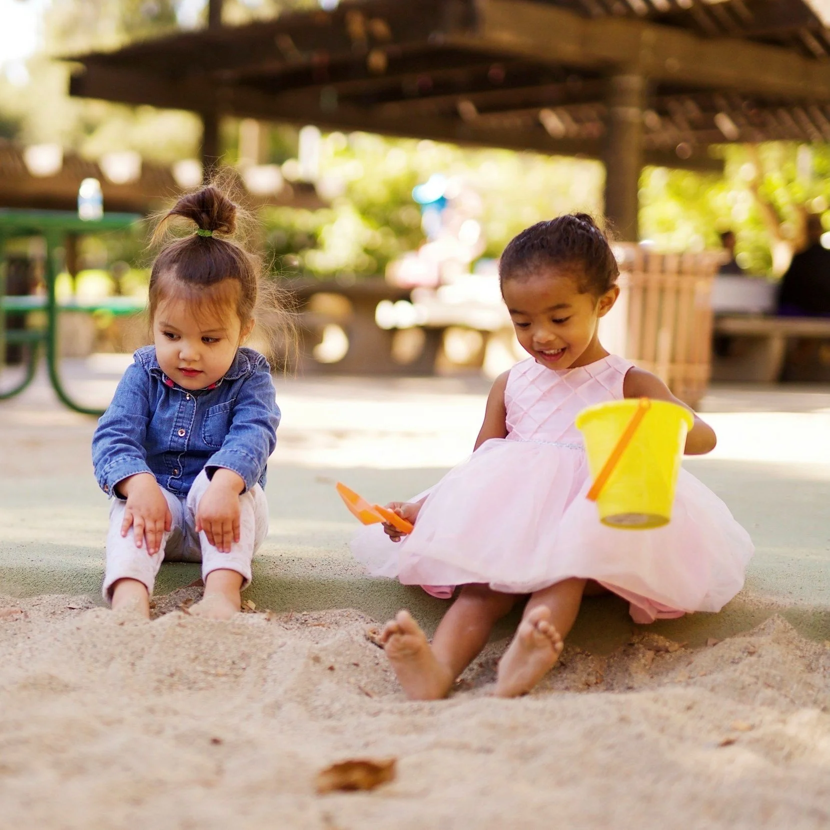 Two young girls playing in a sandbox at a park, one in a pink dress holding a yellow bucket and the other in a blue shirt and white pants.