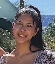 Young woman with dark hair tied back, smiling outdoors, in front of a blue sky and trees.