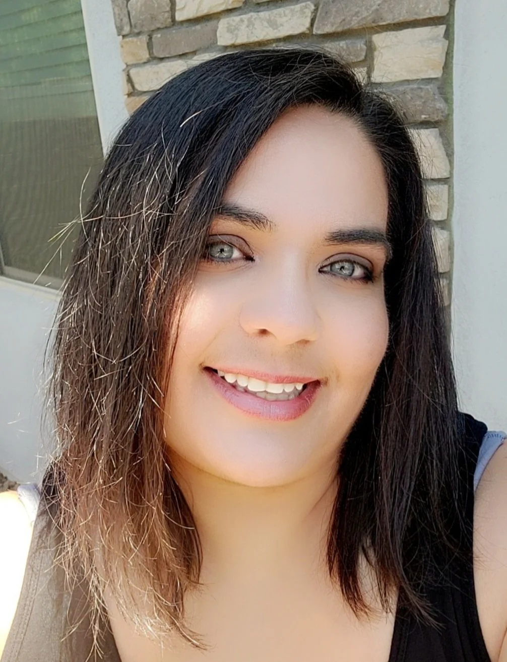 Close-up of a woman with shoulder-length black hair, blue eyes, and a bright smile, standing outside near a house with a stone wall.