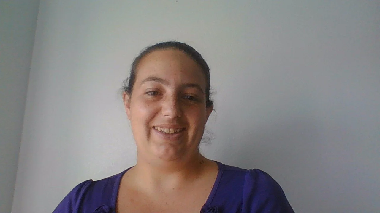 A woman with dark hair in a ponytail smiling at the camera, wearing a purple top.