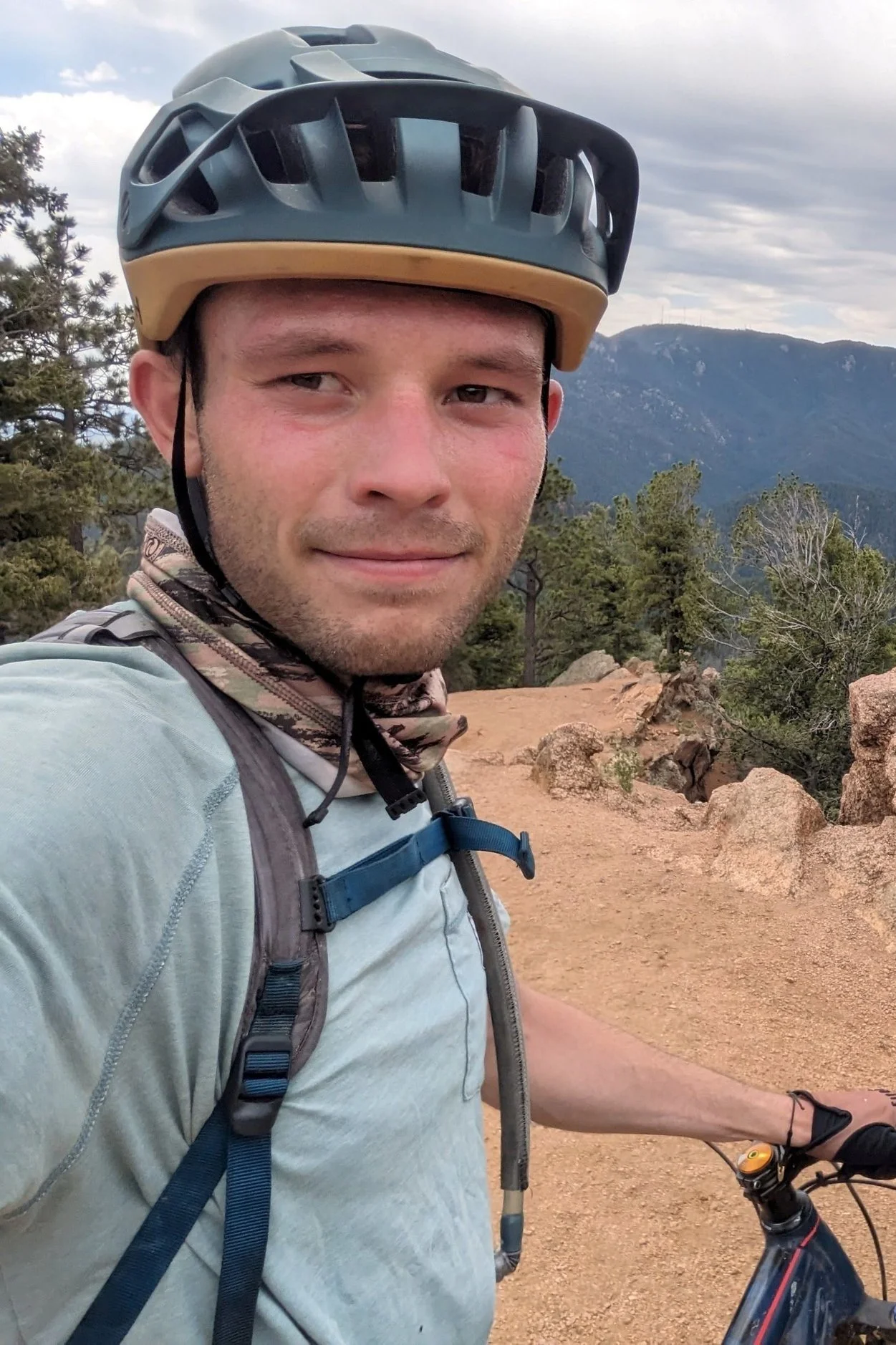 A man wearing a helmet and backpack taking a selfie while mountain biking on a dirt trail with trees and mountains in the background.