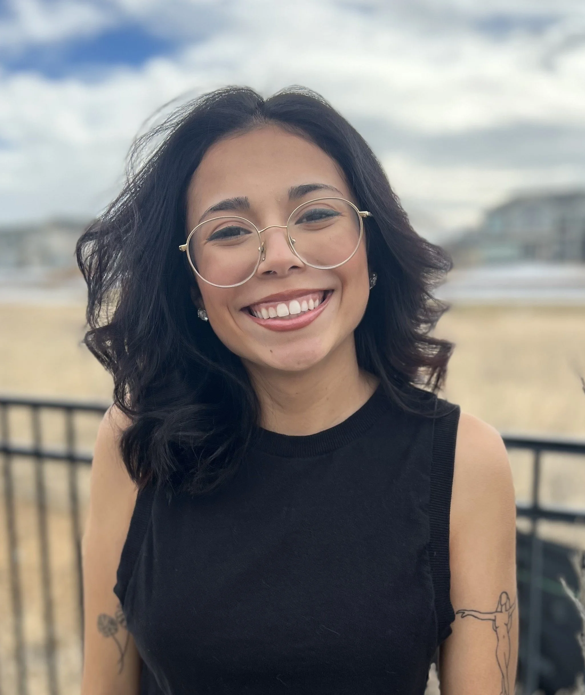 A smiling woman with black wavy hair, wearing glasses and a sleeveless black top, standing outdoors on a cloudy day with a beach and buildings in the background.