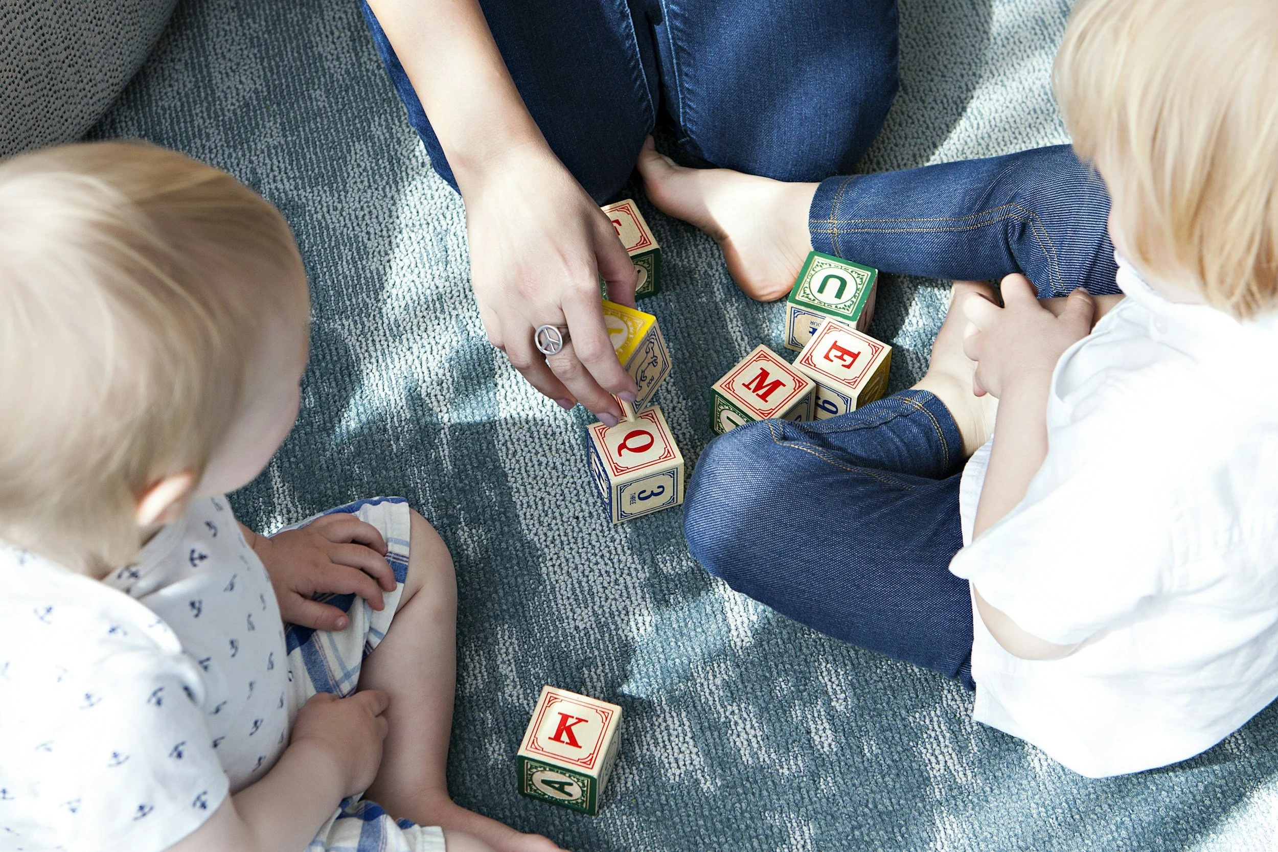 Two children with light hair and an adult sitting on a blue carpeted floor, playing with colorful alphabet blocks. The adult is arranging the blocks while the children watch.