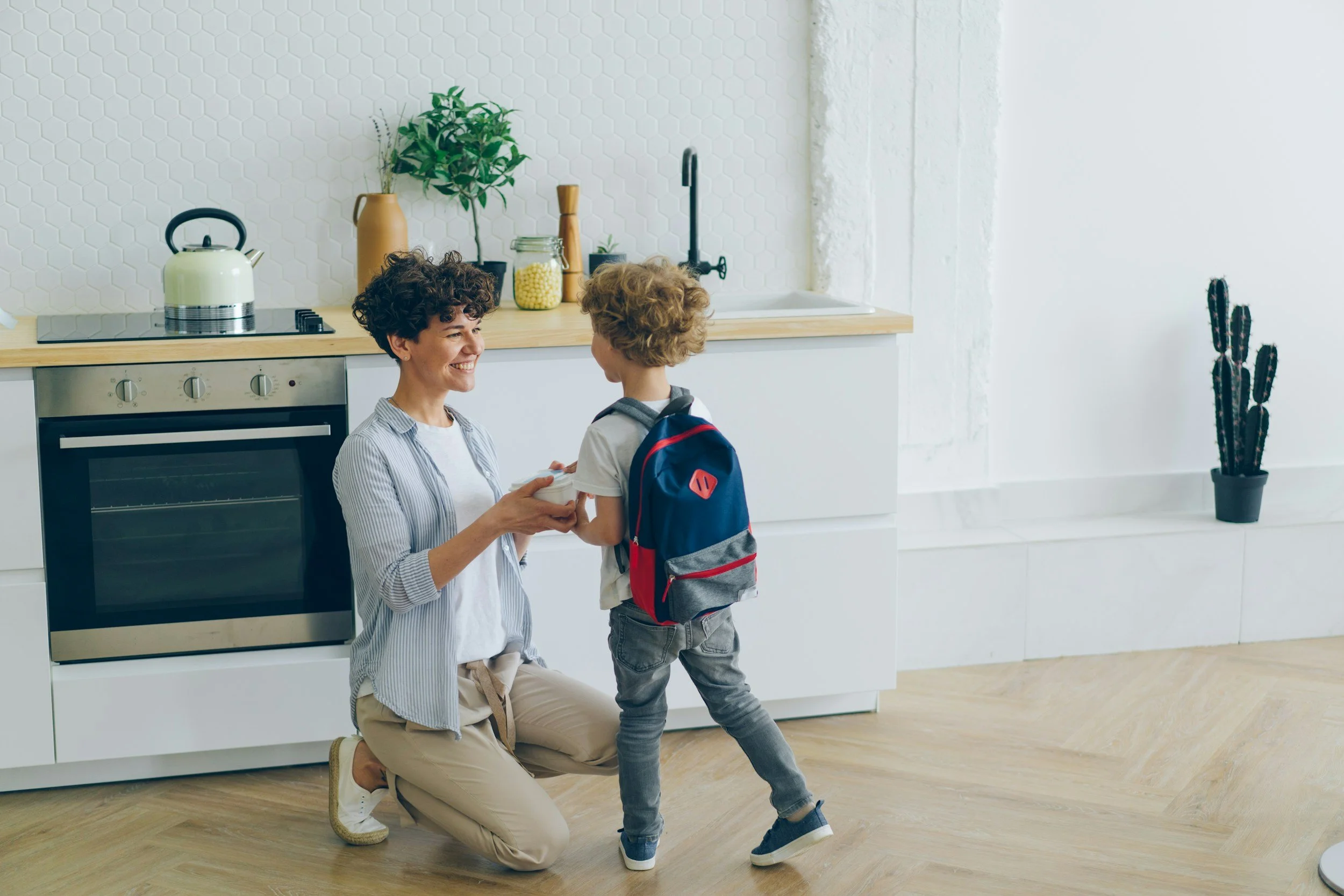 A woman kneeling in a kitchen, smiling and giving a cup to a young boy with a backpack.