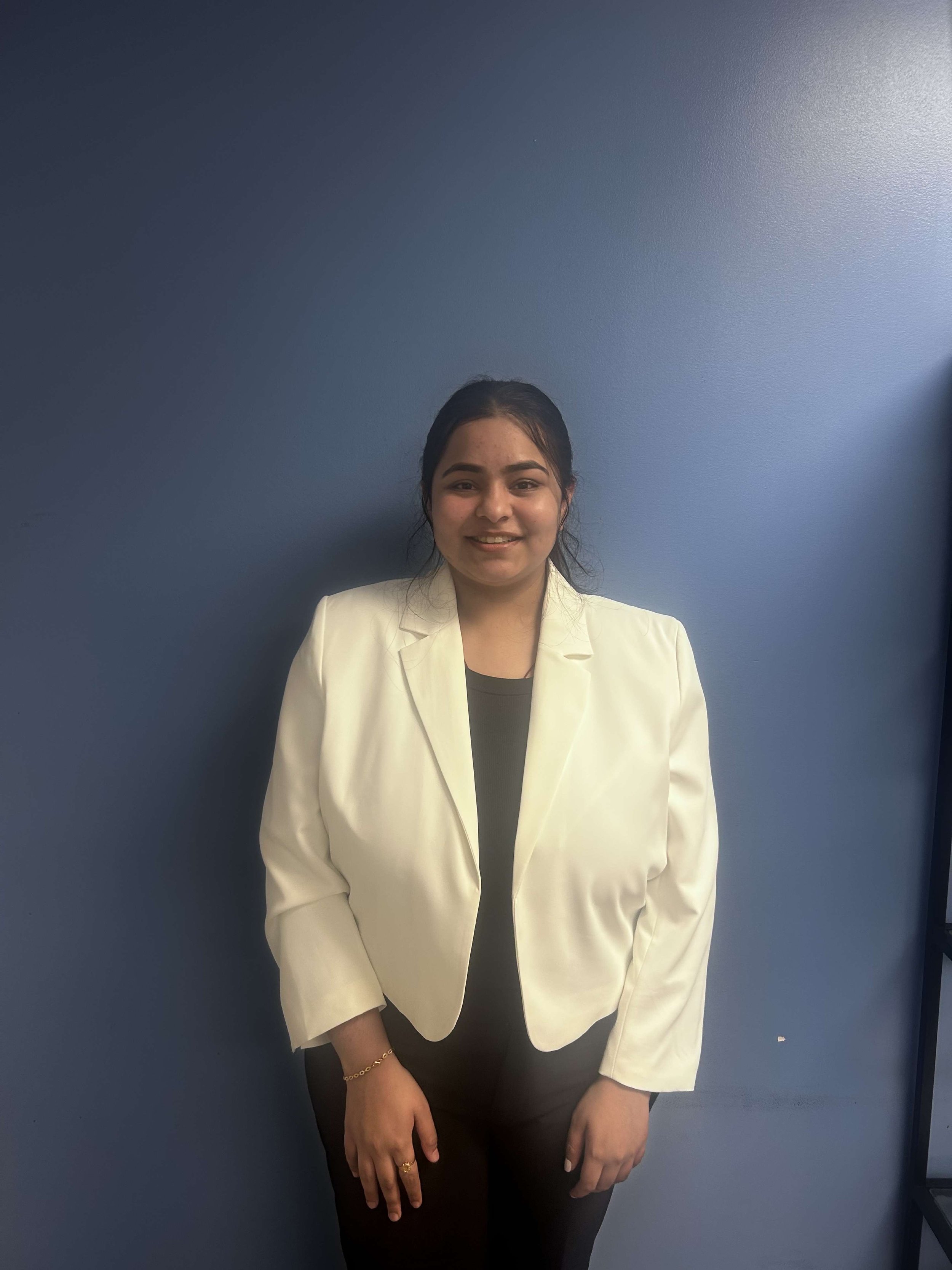 Young woman with dark hair wearing a white blazer and black top standing against a plain blue wall.
