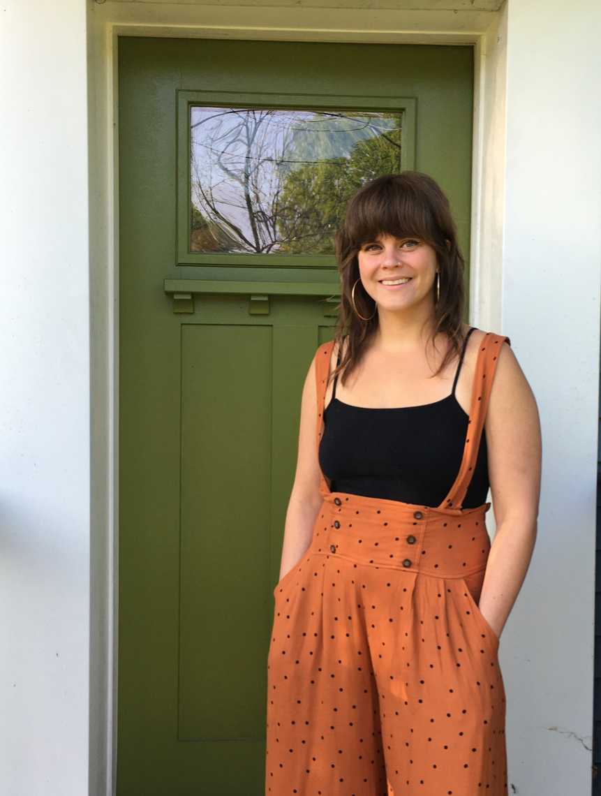 A woman with shoulder-length brown hair and hoop earrings standing outside in front of a green door with a window. She is wearing a black tank top and high-waisted orange pants with black polka dots. She is smiling with one hand in her pocket.