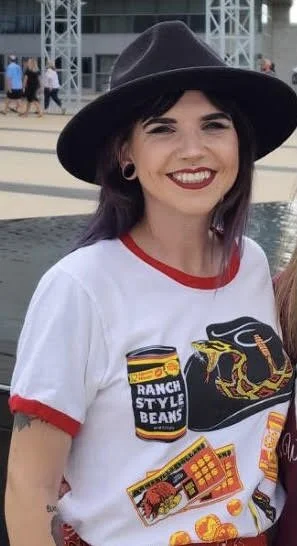 A young woman smiling, wearing a black wide-brimmed hat and a white t-shirt with red trim and colorful graphic prints, including a can of ranch style beans and a snake illustration, standing outdoors near water with other people in the background.