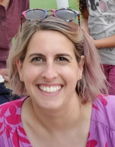 A smiling woman with short blonde hair, wearing glasses on her head and a pink and purple patterned shirt, outdoors with children in the background.