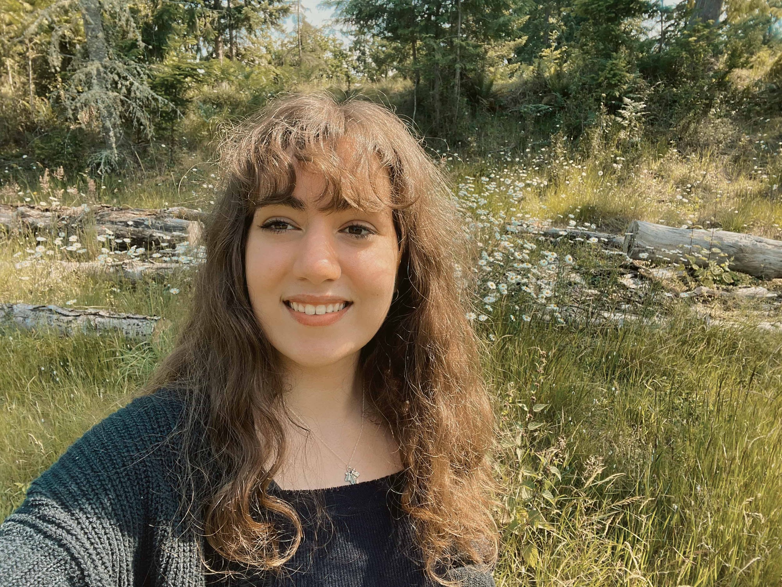 A young woman with light brown, curly hair and fair skin is smiling in a natural outdoor setting with green grass, white wildflowers, and trees in the background.