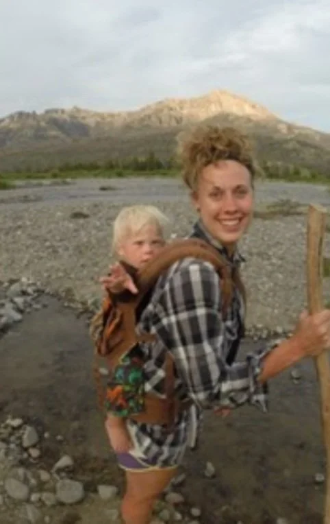 Woman smiling while hiking with a child in a backpack carrier on her back, holding a walking stick, in a rocky outdoor landscape with mountains in the background.