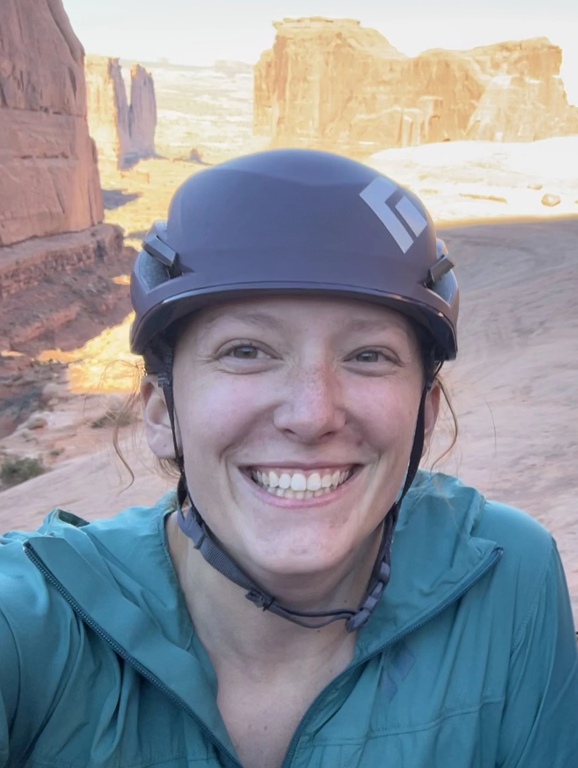 A woman with a helmet smiling outdoors with red rock formations and a desert landscape in the background.