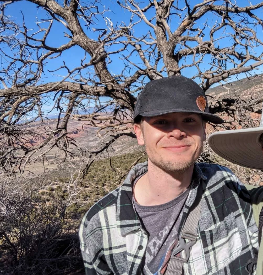 A smiling man in a black cap and plaid shirt taking a selfie outdoors with a leafless tree and desert landscape in the background.
