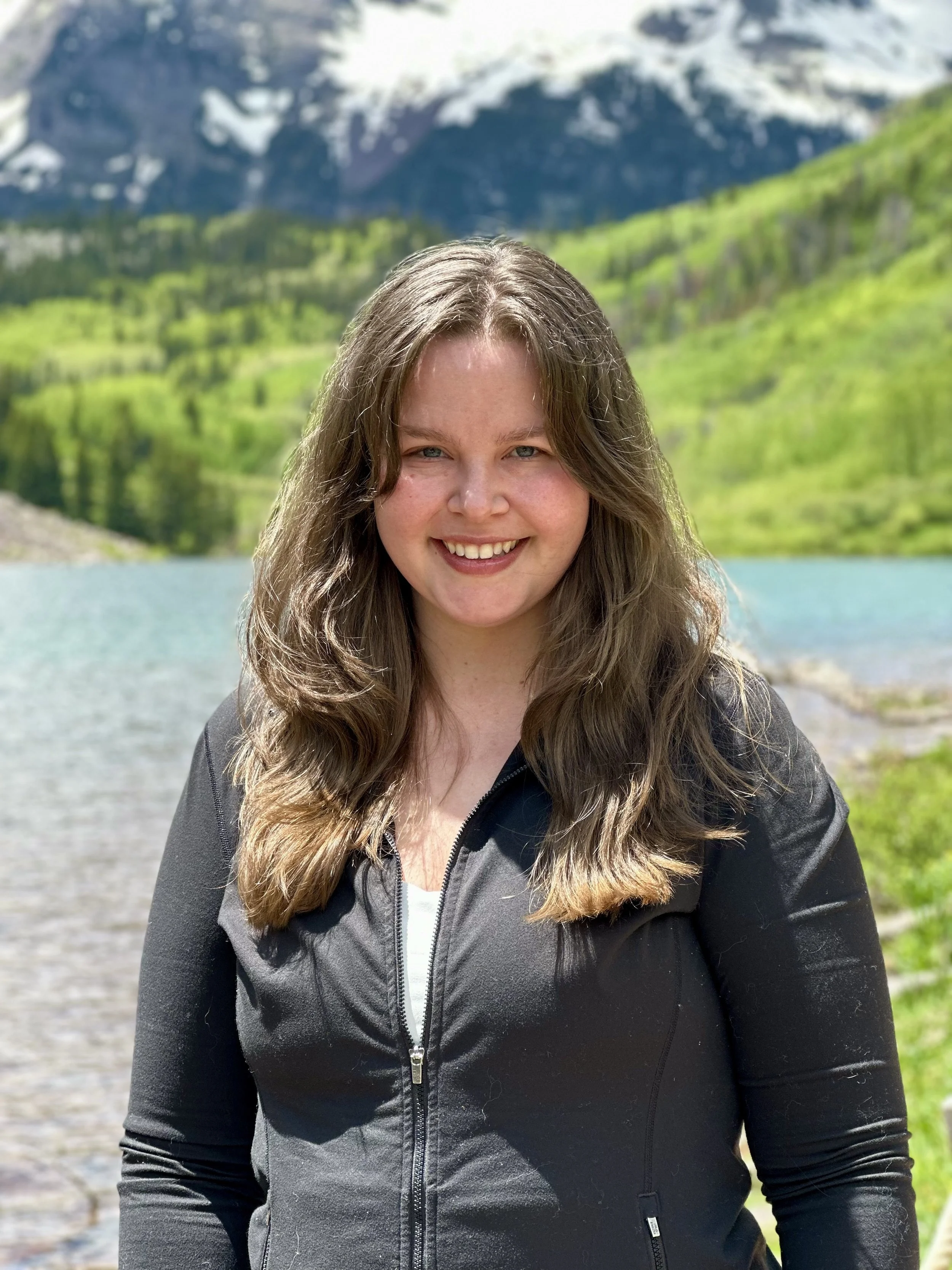 A smiling woman standing outdoors near a lake, with green hills and snow-capped mountains in the background.