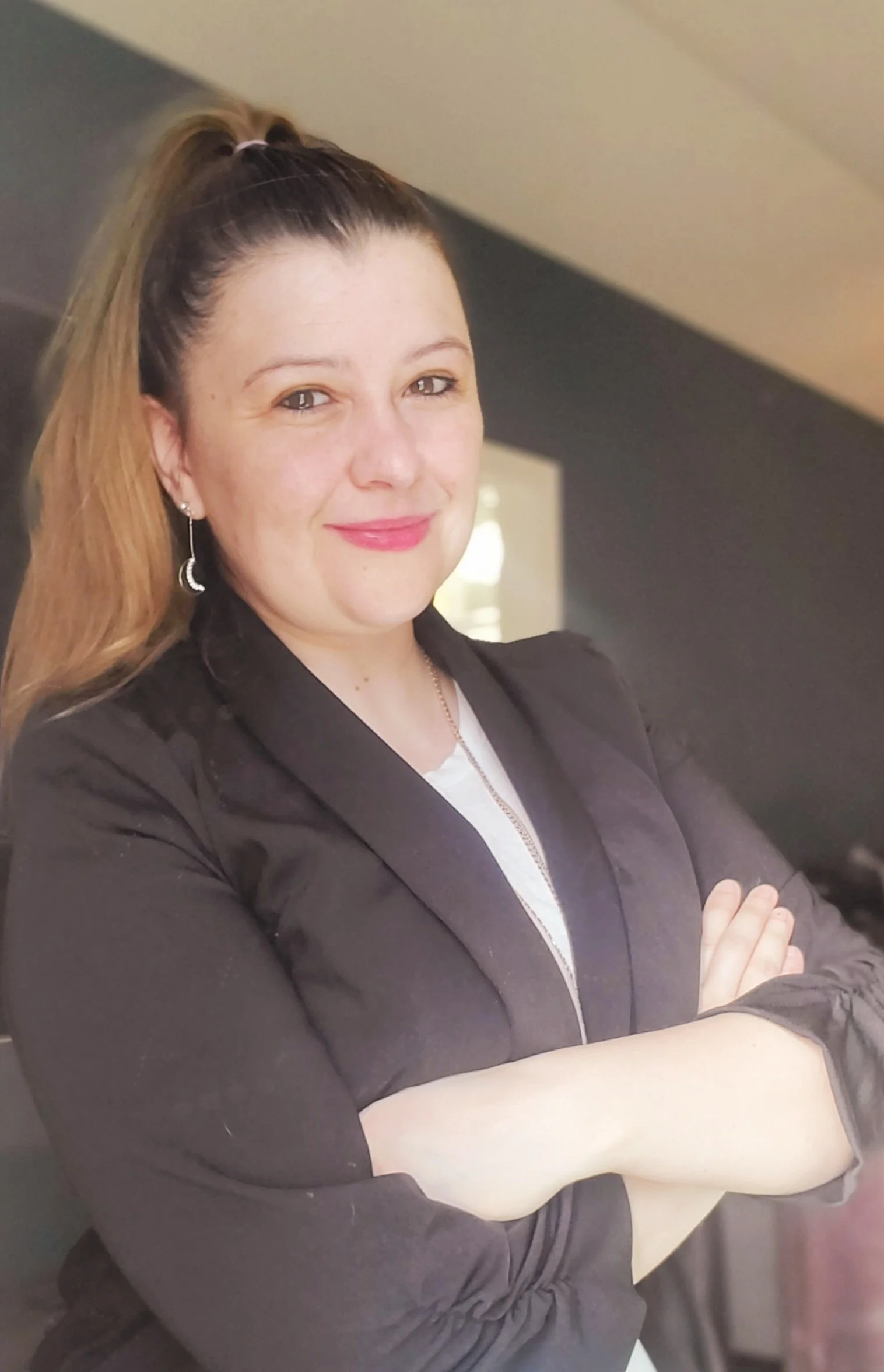 A woman with light skin, long brown hair in a high ponytail, wearing a black blazer, white top, earrings, and a necklace, standing with arms crossed in an indoor setting.