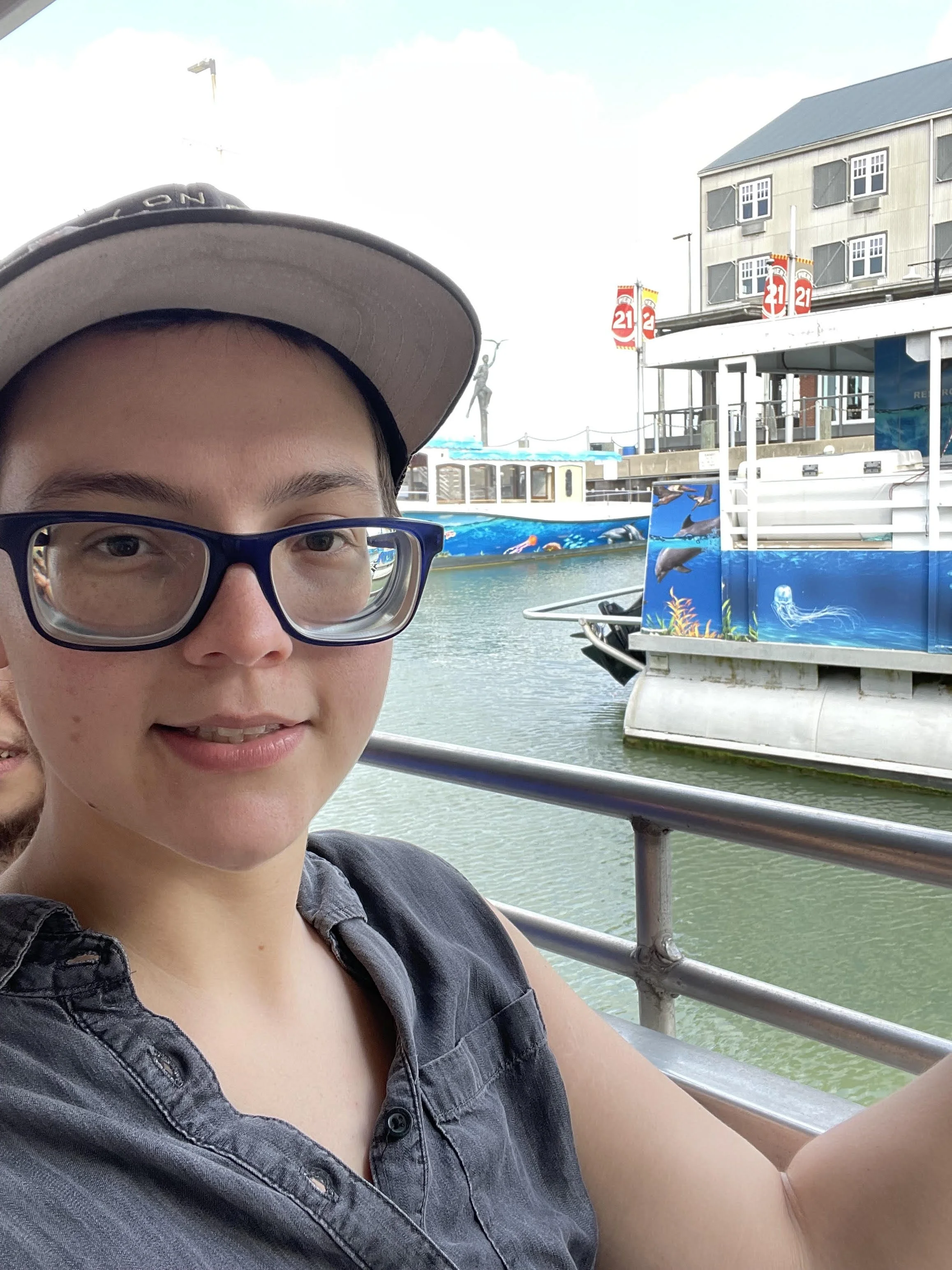 A person taking a selfie on a boat at a waterway, with buildings, a statue, and a painted boat in the background.