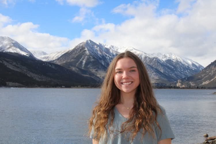 A young woman with long wavy hair smiling in front of a lake, with snow-capped mountains in the background.