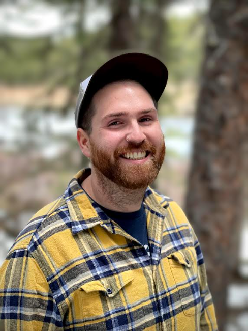 A smiling man with a beard wearing a yellow and blue plaid shirt, a dark shirt underneath, and a baseball cap outdoors with trees and water in the background.