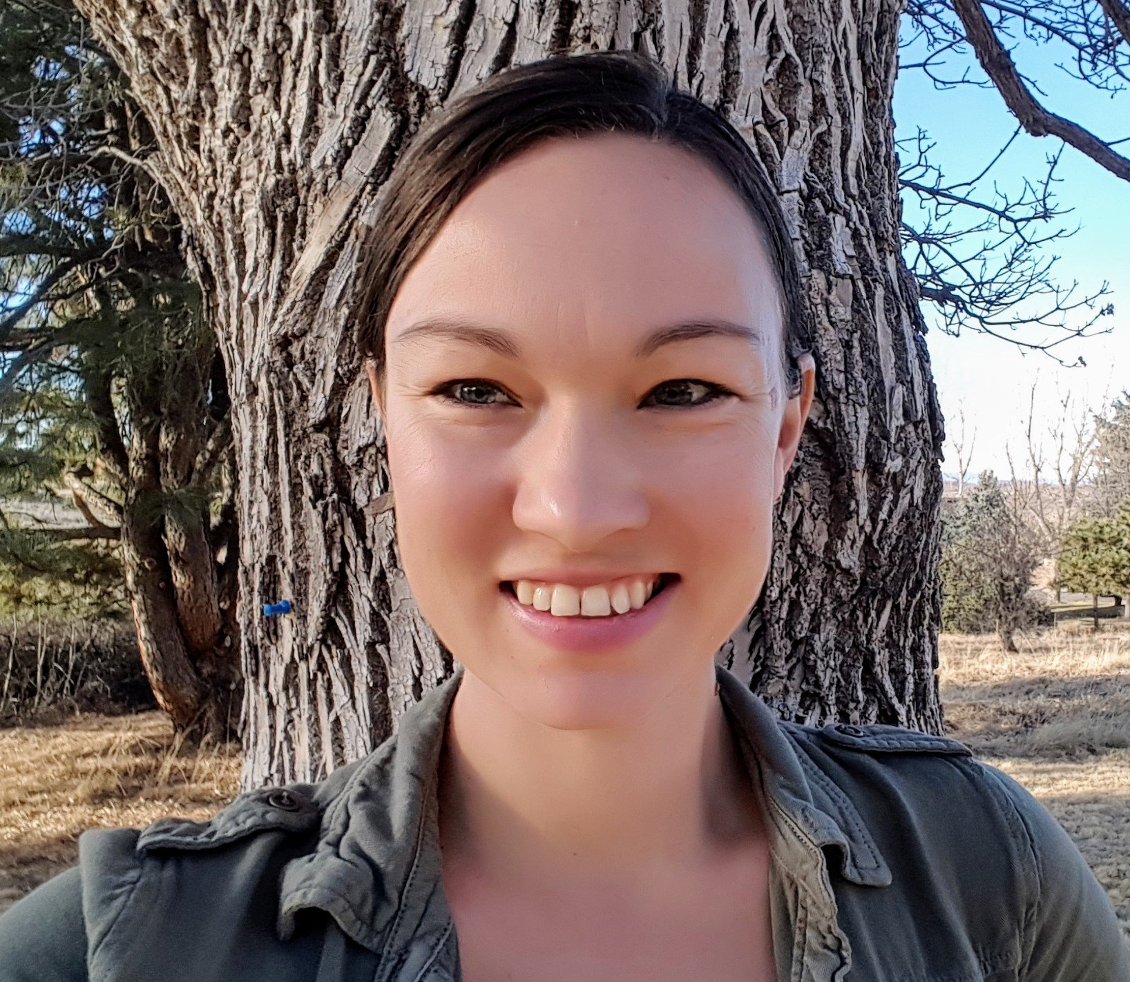 A smiling woman with short dark hair standing in front of a large tree with textured bark, outdoors on a sunny day with clear sky and some distant trees.