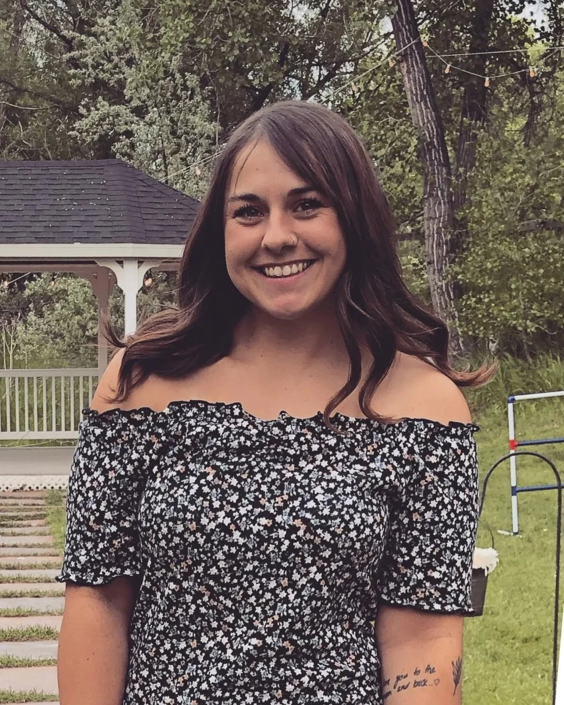 A young woman with dark brown hair smiling outdoors in front of a garden and gazebo, wearing an off-the-shoulder black dress with white floral pattern.