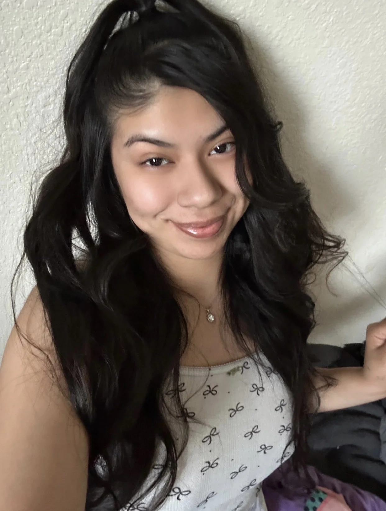 A woman with long, dark, curly hair smiling at the camera, wearing a white top with a pattern of small black bows, gold necklace, and standing against a beige wall.
