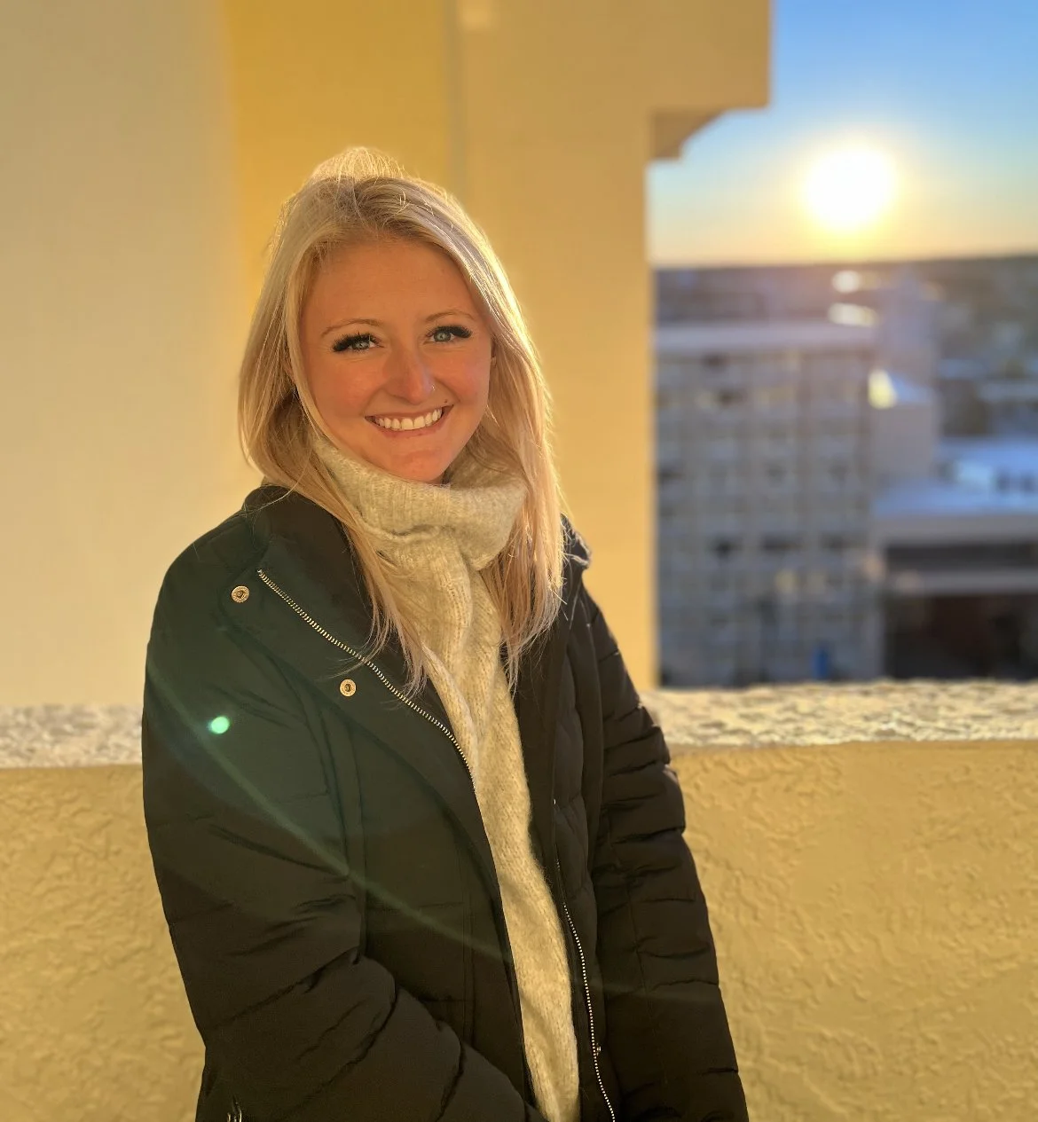 A smiling woman with blonde hair wearing a black jacket and a tan turtleneck sweater standing on a balcony at sunset, with city buildings and the sun in the background.